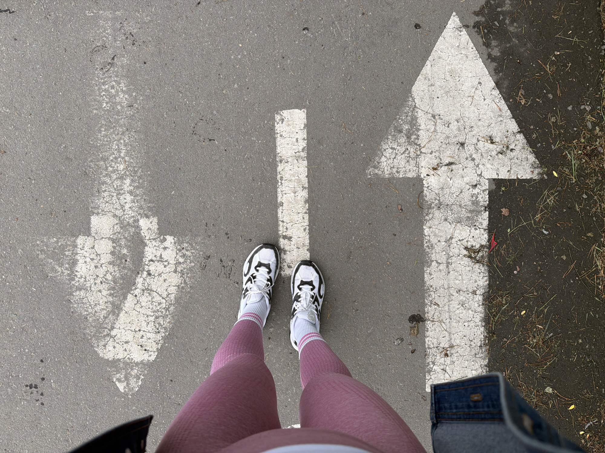 An overhead, high-angle medium shot shows a person's legs and feet wearing pink leggings, white socks with pink stripes, and white and black athletic shoes standing on a gray asphalt surface. Three white directional arrows are painted on the asphalt. The closest arrow points to the right, and is mostly within the frame, partially obscured by the person's feet. A second arrow, also pointing right, is positioned above the first and is partially cut off by the top of the frame. The third arrow, pointing to the left, is positioned above the second, and is also partially cut off by the top of the frame. The asphalt is cracked and worn in places. There is some debris on the asphalt and to the right of the closest arrow, including dry leaves, twigs, and what appears to be animal droppings. The lower left corner of the image shows the edge of a person's denim garment. The scene appears to be outdoors, possibly on a sidewalk or road. The location is the city of Birobidzhan, Russia.