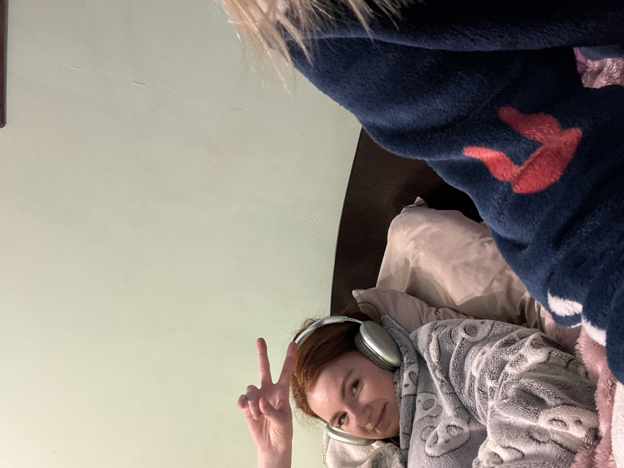 A bedroom scene featuring a person lying in bed, angled with the head on the right side of the frame. The person has red hair, is wearing silver headphones, and is covered with a gray blanket featuring a pattern of faces. They are making a peace sign with their left hand. An object covered with a blue blanket with a red design is partially visible in the upper right corner. The wall is light green. A headboard is visible.