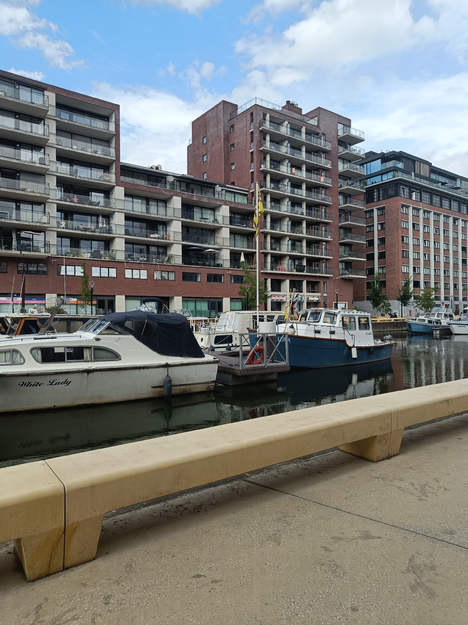 The image shows a canal-side view of modern apartment buildings in Leuven, Belgium. In the foreground, a concrete bench sits on a paved walkway, with the canal water reflecting the sky and buildings. Several boats are docked along the canal. The closest boat, a white vessel named "White Lady," has a dark blue cover over its cockpit. Behind it, a blue and white boat with the name "LUDA" visible is also docked. Further down the canal, more boats and apartment buildings are visible. The buildings are constructed with red brick and feature balconies with glass railings. The sky is partly cloudy.