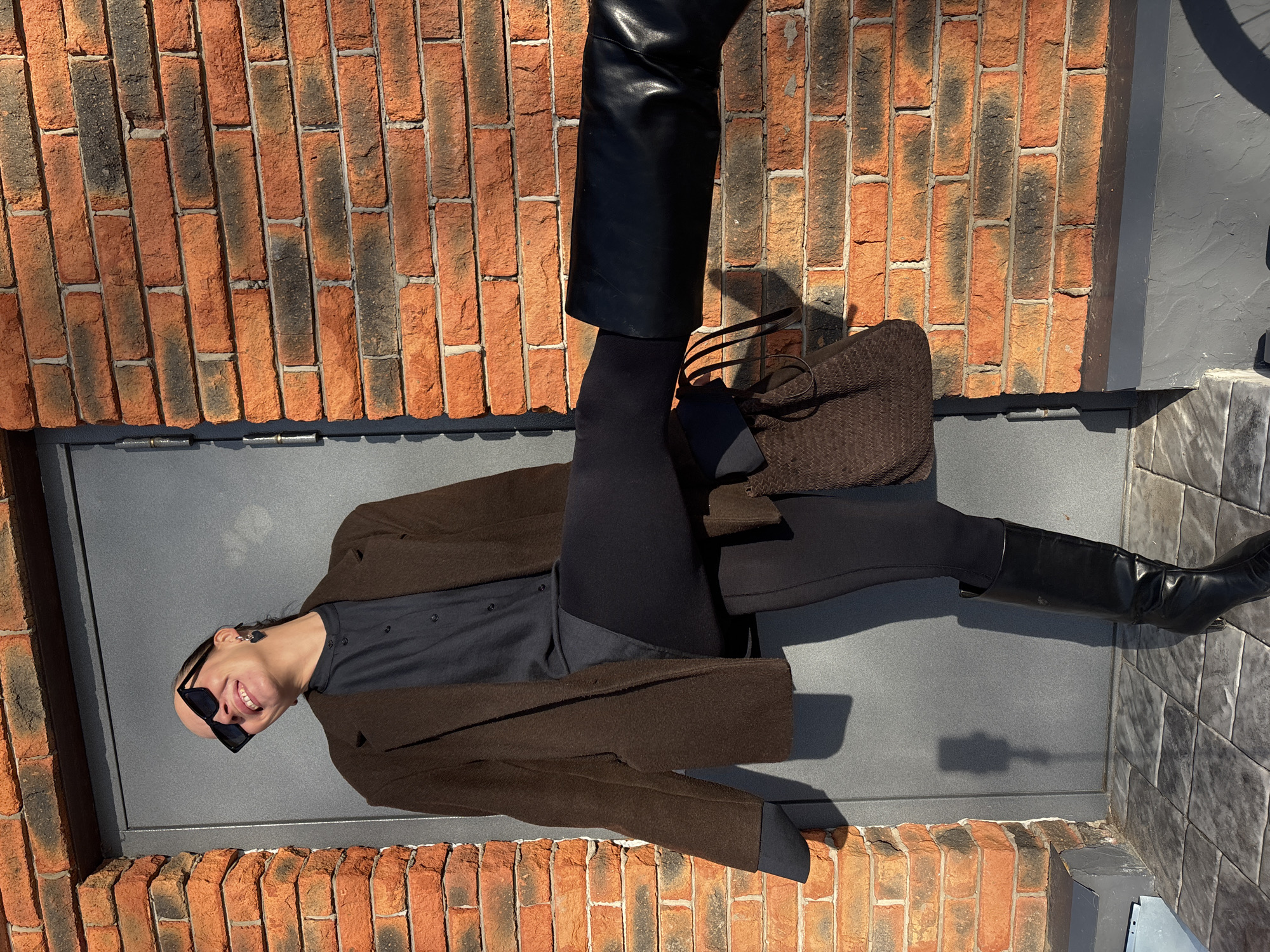A person is lying on a gray, possibly metal, surface between two brick walls in what may be an urban environment. The person is wearing sunglasses, a dark shirt, a brown coat, black tights, a black leather skirt, and black boots. A brown bag is next to them. The environment has both red brick and a tile wall. The image is not oriented in a way that suggests standard perspective, implying an unusual camera angle or a manipulated image. There is no immediately verifiable evidence that connects this scene to the city of Chaikovsky, Russia.