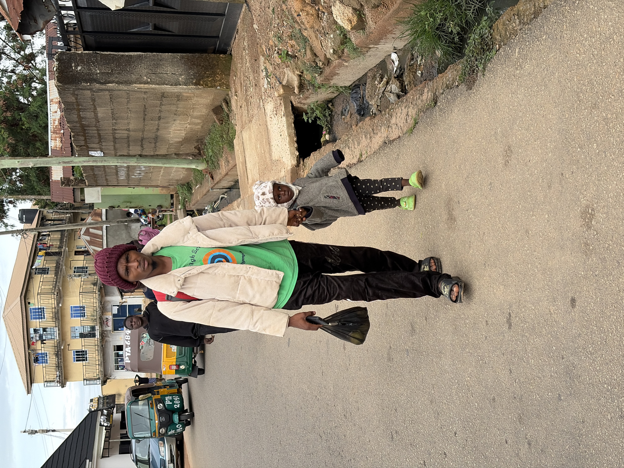 In Jos, Nigeria, a man and a young child walk hand-in-hand on a paved road. The man, wearing a maroon beanie and a cream-colored jacket over a green t-shirt, carries a black plastic bag. He has dark pants and sandals on his feet. The child, dressed in a grey jacket, patterned leggings, and bright green shoes, also wears a white hat with ear-like protrusions. They are walking alongside a concrete drainage ditch filled with debris. In the background, several buildings are visible, including a multi-story yellow building with balconies and laundry hanging out. To the left of the road, a light blue car and two auto-rickshaws (Keke Napep) are parked or moving. A brick wall runs along the right side of the road, with a metal gate at its end. The sky is overcast.