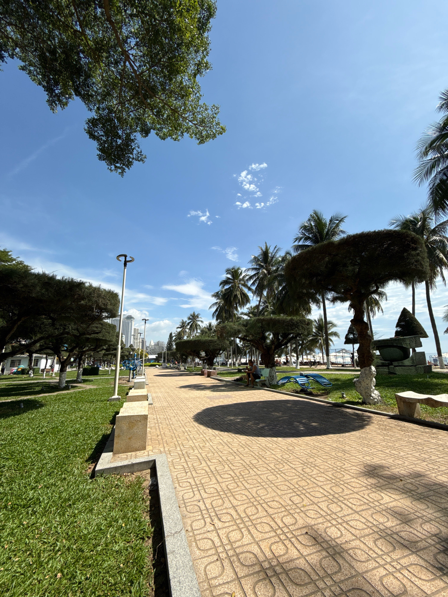 This is a photograph of a park with a paved walkway in Nha Trang, Vietnam. The walkway is paved with light-colored square tiles with a decorative pattern. To the left of the walkway is a well-maintained lawn. Further to the left, several trees with lush green foliage are visible, as well as lampposts. In the background, there are tall buildings, palm trees, and beach umbrellas, suggesting proximity to a beach. A few people are seated on benches and some exercise equipment is visible in the park. The sky is blue with scattered white clouds, and the sun is casting shadows on the walkway.