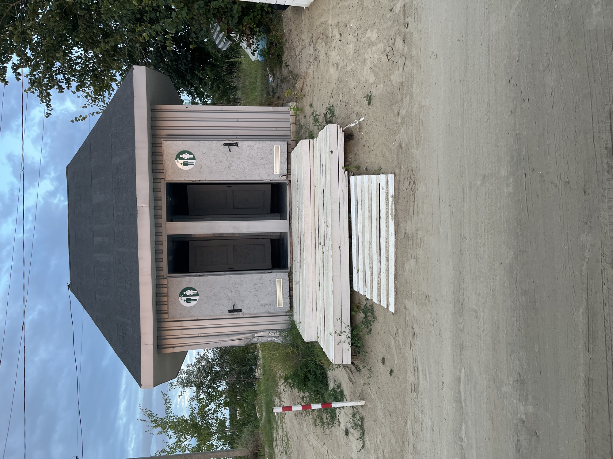 The image shows a pair of public toilets located on a sandy area in Bezliudivka, Ukraine. The structure is small and appears to be made of metal siding with a dark grey, gabled roof. Each of the two identical doors is marked with a circular green and white sign depicting male and female figures, indicating separate facilities for men and women. The toilets are elevated on a wooden platform made of light-colored planks. A red and white striped pole stands to the left of the entrance, and vegetation grows around the base of the structure and on the sandy ground. In the background, trees and a cloudy sky are visible. The ground in front of the toilets is a dirt road with tire tracks, suggesting it is a well-trafficked area. There are no people visible in the image.