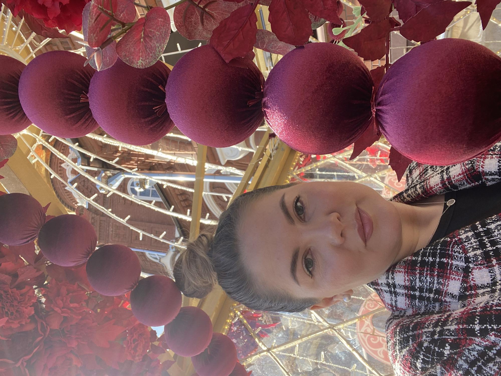The image shows a woman with her hair in a bun, wearing a black top and a red and black patterned jacket. She is positioned beneath a decorative ceiling featuring rows of maroon velvet spheres resembling Christmas ornaments, interspersed with red flowers and leaves. The spheres are suspended from a structure of white metal bars. The background reveals a building with architectural details visible through a glass and metal framework, suggesting an interior space. The overall setting appears to be festive and ornamental. Based on the details, the location appears to be the Central Square of Izhevsk, Russia, known for its decorative installations, particularly during holiday seasons.