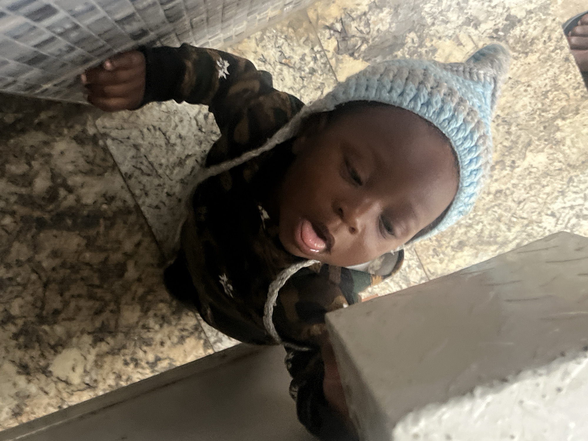 A baby wearing a blue, knit hat and a patterned shirt is standing in what appears to be a bathroom or kitchen. The baby is near a counter or cabinet, and part of a person's foot is visible in the upper right corner of the image. The location is Gura Top, Nigeria.