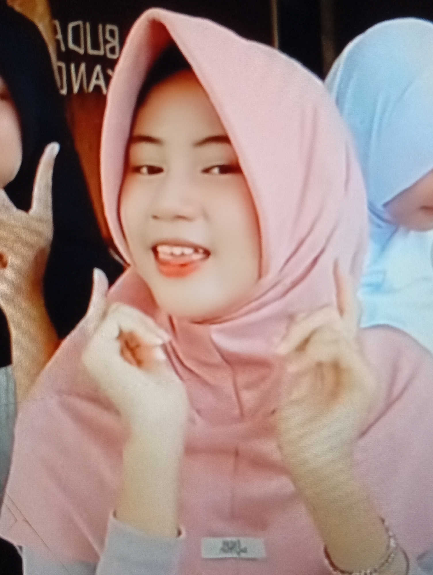 The image is a close-up, medium-shot photograph of a young woman wearing a light pink hijab. Her face is turned slightly towards the viewer, and she is smiling with her mouth open, revealing her teeth. Her eyes are looking forward, and she is making a gesture with her right hand, with her index finger and thumb forming a circle. She is wearing a light pink garment that appears to be a t-shirt or blouse. To her left, another person's face is partially visible, wearing a black hijab. To her right, another person is wearing a light blue hijab, only the head and shoulder are visible. The background is dark and out of focus, with some blurred text that appears to read "BUDU" and "YANG" in white. The image quality is low, with visible pixelation and a warm color cast. The specific location in the city of Lhokseumawe, Indonesia, cannot be determined from the provided image.