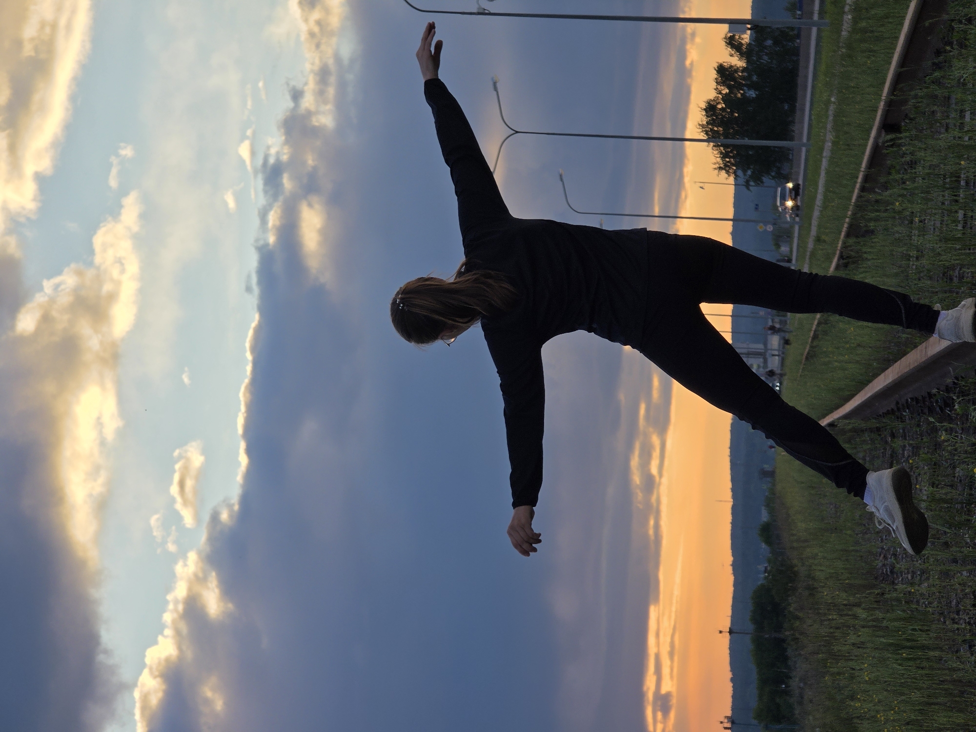 A female figure, back to the camera, is poised precariously on a railway track against a sunset sky in Chaikovsky, Russia. She is dressed in black workout gear and white sneakers, her arms outstretched, one reaching up towards the sky and the other down towards the train track. Her dark hair is pinned back. The sky is divided into zones of blue and orange, suggesting either sunrise or sunset. Lampposts are visible in the background, as well as trees and a road with car headlights illuminating the scene. The railway track is overgrown with green vegetation.