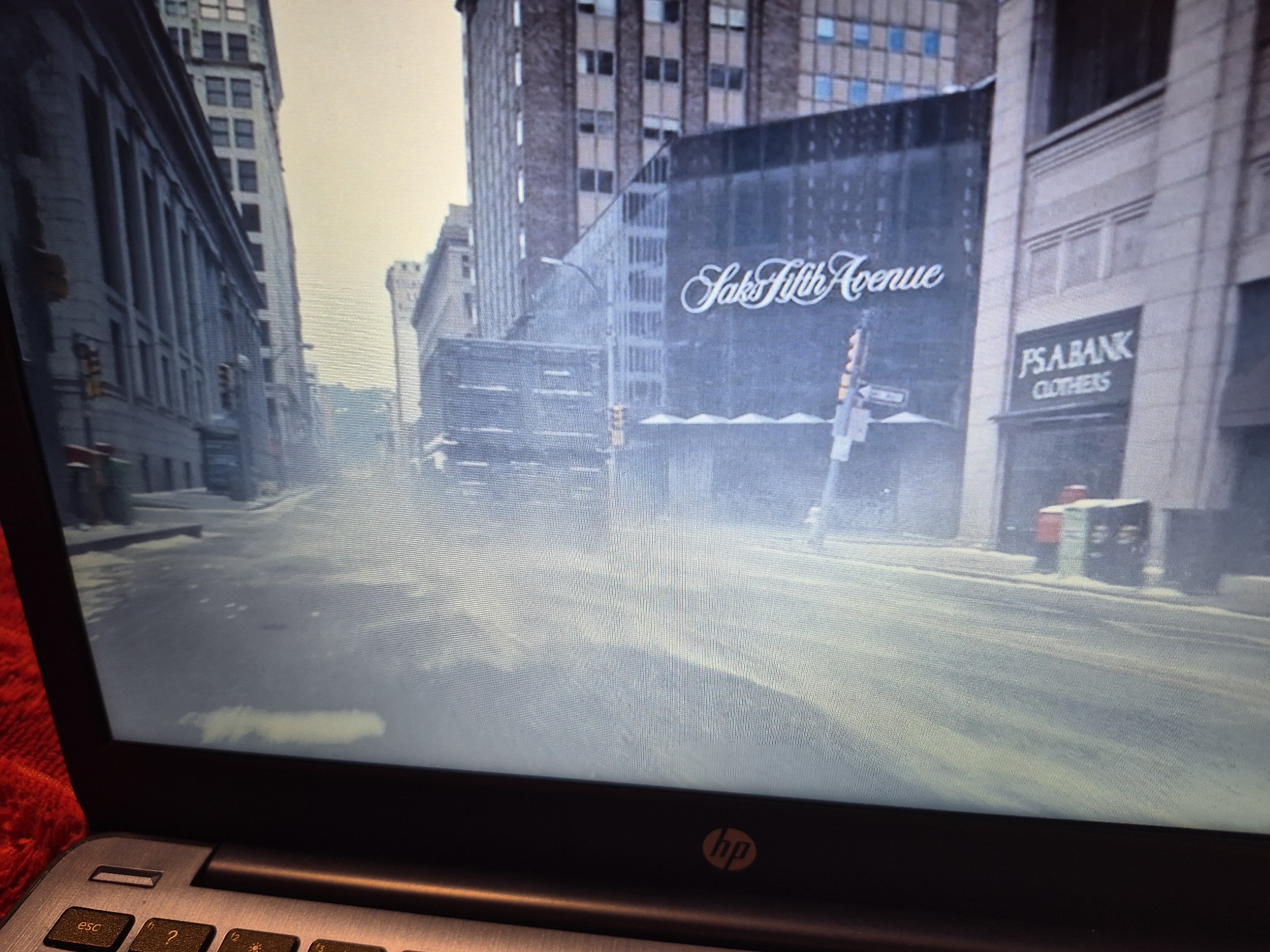 The image shows a street scene from a video game, possibly Grand Theft Auto IV, given the aesthetic and the presence of recognizable landmarks. The scene depicts a city street with tall buildings lining both sides. In the center, a Saks Fifth Avenue store is visible with its distinctive signage. To the right, a PS A BANK CLOTHERS store is also identifiable. The street appears to be wet or covered in mist, giving it a hazy and somewhat desolate atmosphere. There are traffic lights and street signs visible. The foreground shows a portion of a laptop keyboard, suggesting the image was captured from a screen. There are no people or animals depicted in the scene. The overall environment appears to be urban, with a focus on architecture and commercial establishments. The location is not in Sandwell, United Kingdom; it is a depiction of a fictionalized New York City, specifically Fifth Avenue.