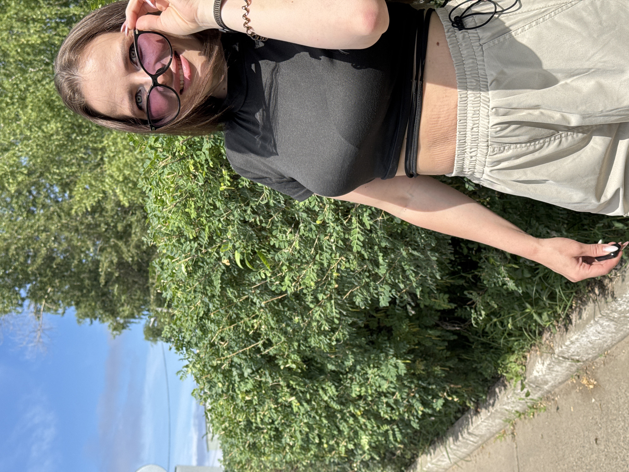 A young woman with brown hair stands in front of a dense green hedge with a sidewalk and curb to her left, under a bright blue sky with scattered clouds. She is wearing a black crop top, light beige cargo pants, and sunglasses. Her right hand is raised to adjust her sunglasses, and her left arm is bent at the elbow. The scene appears to be a sunny day in a residential area of Chaikovsky, Russia, likely on a street or path lined with trees and hedges.