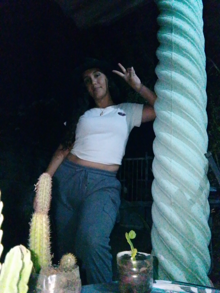 This image depicts a female individual posing in a dimly lit outdoor or semi-outdoor setting in Juan Griego, Venezuela. The subject, an adult with long, dark, wavy hair, is wearing a dark hat, a white short-sleeved crop top, and dark gray cargo sweatpants. She is positioned towards the center-left, with her right arm leaning against a prominent light-colored, spiraled column on the right side of the frame. Her left hand is raised, forming a "peace" sign. She is wearing a necklace with a small pendant and is looking directly at the camera with a slight smile. In the foreground on the left, two potted cacti are visible in clear plastic containers. On the lower right, a small green plant sprout grows in a similar clear container. The background is dark and indistinct, suggesting nighttime, with faint details of foliage and a metallic gate structure discernible behind the subject's lower body.