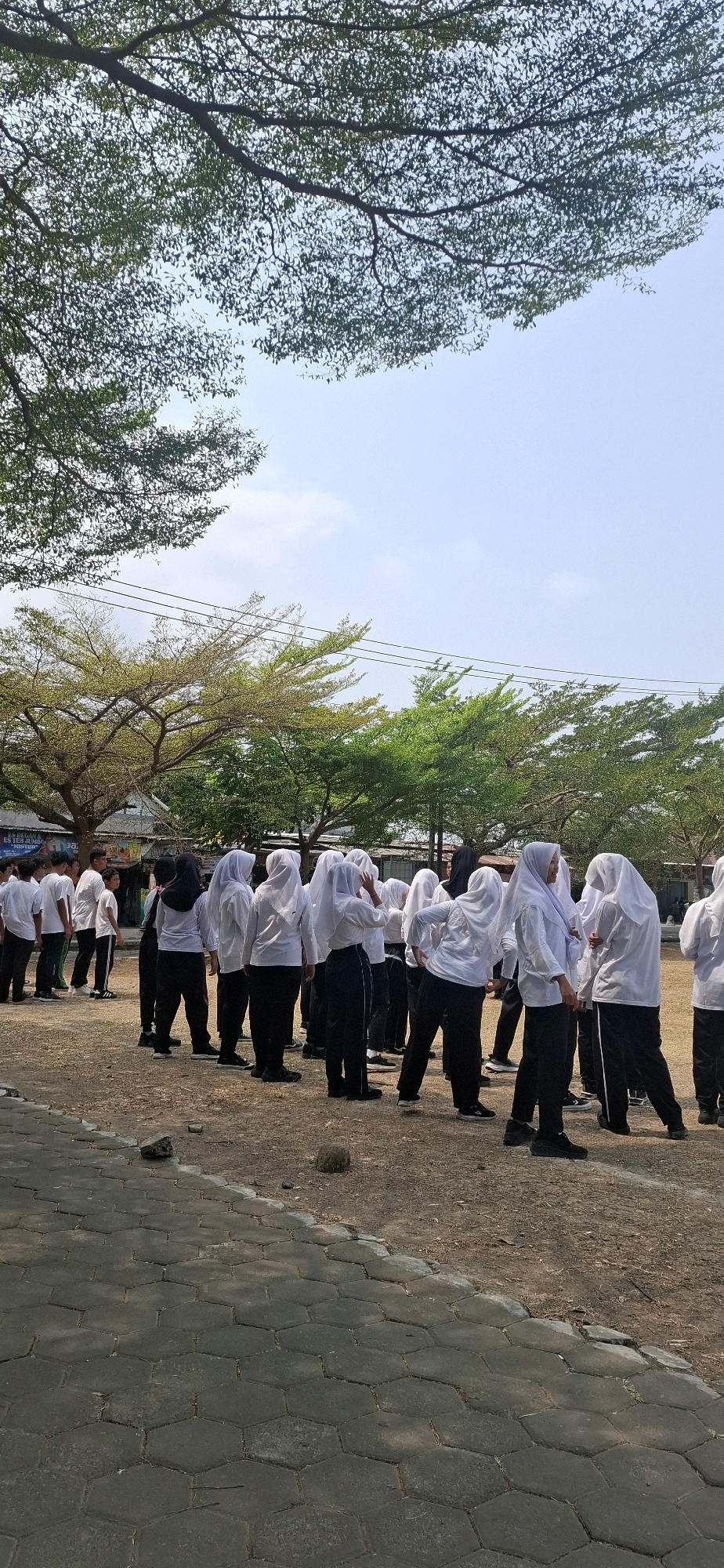 In Baturan, Indonesia, a group of students in white shirts and black pants, many wearing headscarves, stand in a line outdoors on a dirt field. They appear to be participating in a physical activity or formation. The scene is framed by trees with green and yellow foliage, suggesting an outdoor school or community gathering space. The sky is clear with a few clouds. The ground is covered with dry grass and some hexagonal paving stones are visible in the foreground.