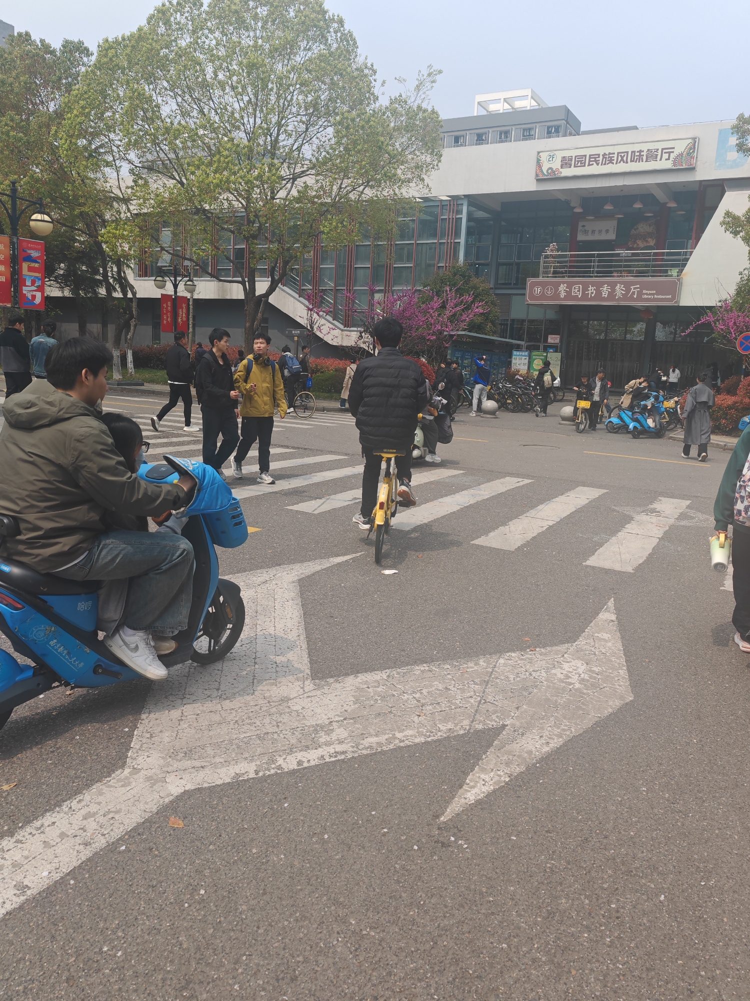 Here is a detailed description of the image:
**Scene Overview:**
The photo appears to be taken at a street intersection or pedestrian crossing in Jiangning District, China. Pedestrians and cyclists are present. The scene appears to be taken during daytime.
**People:**
* There are several people, most notably two people on a blue electric scooter.
* A man is riding a yellow bicycle across the crosswalk.
* There are other pedestrians walking and standing around.
**Objects:**
* Electric scooter with a blue body and a passenger.
* Yellow bicycle being ridden.
* White crosswalk markings and a right-turn arrow on the road.
* Building with signage in Chinese that includes the names "馨园民族风味餐厅" (XinYuan Ethnic Restaurant) and "馨园书香餐厅" (XinYuan Library Restaurant).
* Other bicycles and scooters are parked nearby.
**Actions and Interactions:**
* People are walking across the street.
* Someone rides a bicycle across the crosswalk.
* Two people are riding the electric scooter.
**Environment:**
* Urban setting with a building visible in the background.
* Trees with light green foliage are scattered around.
* The weather appears to be overcast.
**Location:**
Based on the license plate information and the name of Nanjing University of Aeronautics and Astronautics (南京航天大學) on the scooter, as well as the restaurant building, the location is likely in the Jiangning District of Nanjing, China near or on the campus of Nanjing University of Aeronautics and Astronautics.