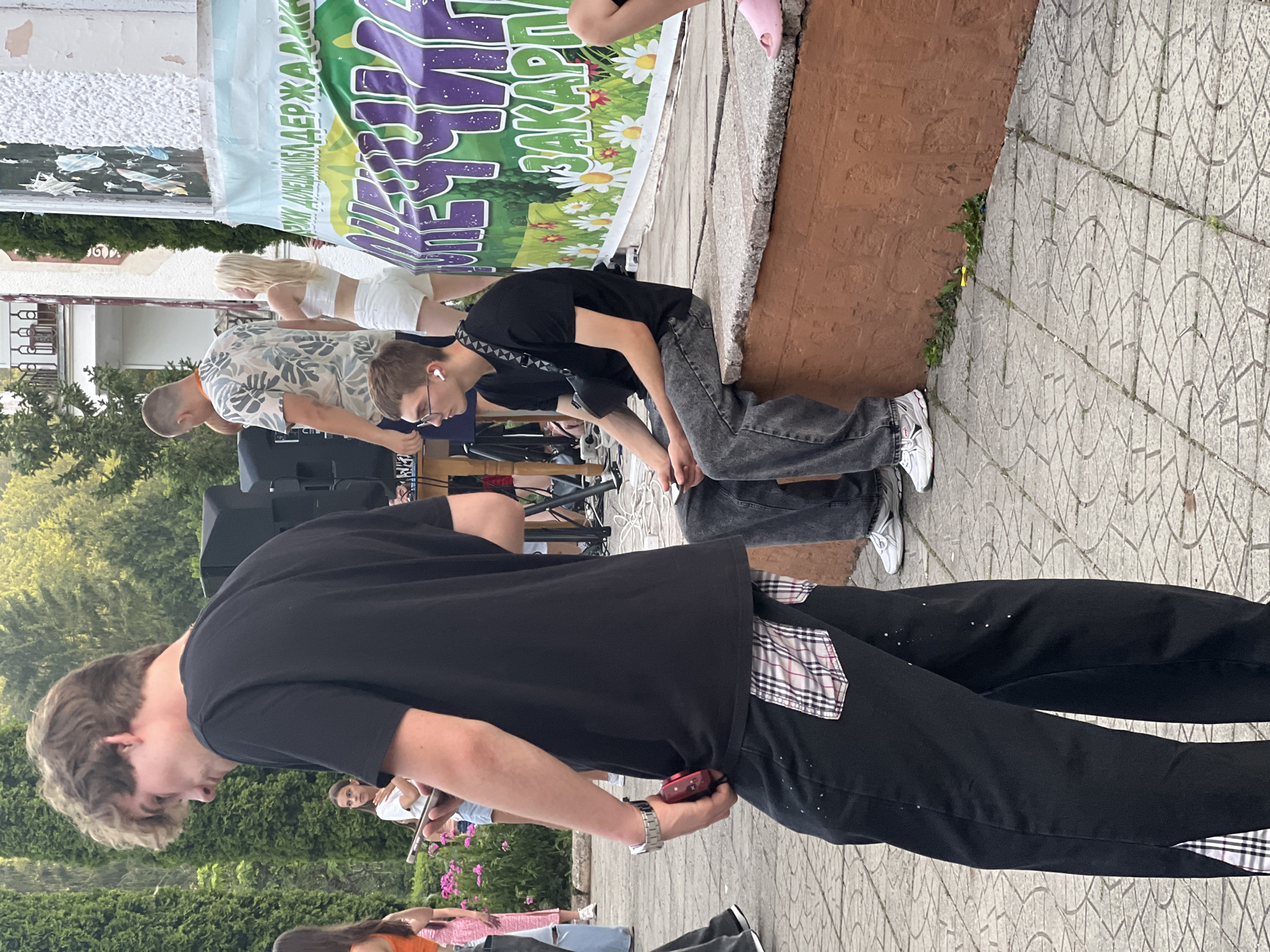 The image depicts an outdoor scene in what appears to be a public area, possibly a park or event space, in the city of Сонячне, Ukraine. In the foreground, a young man with light brown curly hair, wearing a black t-shirt and black pants with a plaid pattern on the back pockets and down the side seams, is captured from the waist down as he walks away from the camera. He is holding a red object in his right hand, tucked into his pocket. To his right, another young man with short brown hair and glasses, wearing a black t-shirt and dark jeans, is seated on a low concrete ledge. He has white wireless earbuds in his ears and a dark bag slung across his chest. He is holding a smartphone in his hands. In the background, other individuals are visible, including a man in a patterned shirt and a blonde woman in a white crop top and shorts standing near a speaker. A large banner with text and floral imagery is displayed behind them. The ground is paved with light-colored square tiles. Green foliage and trees are present in the background, suggesting a summery or spring environment. The overall impression is a casual gathering or event.