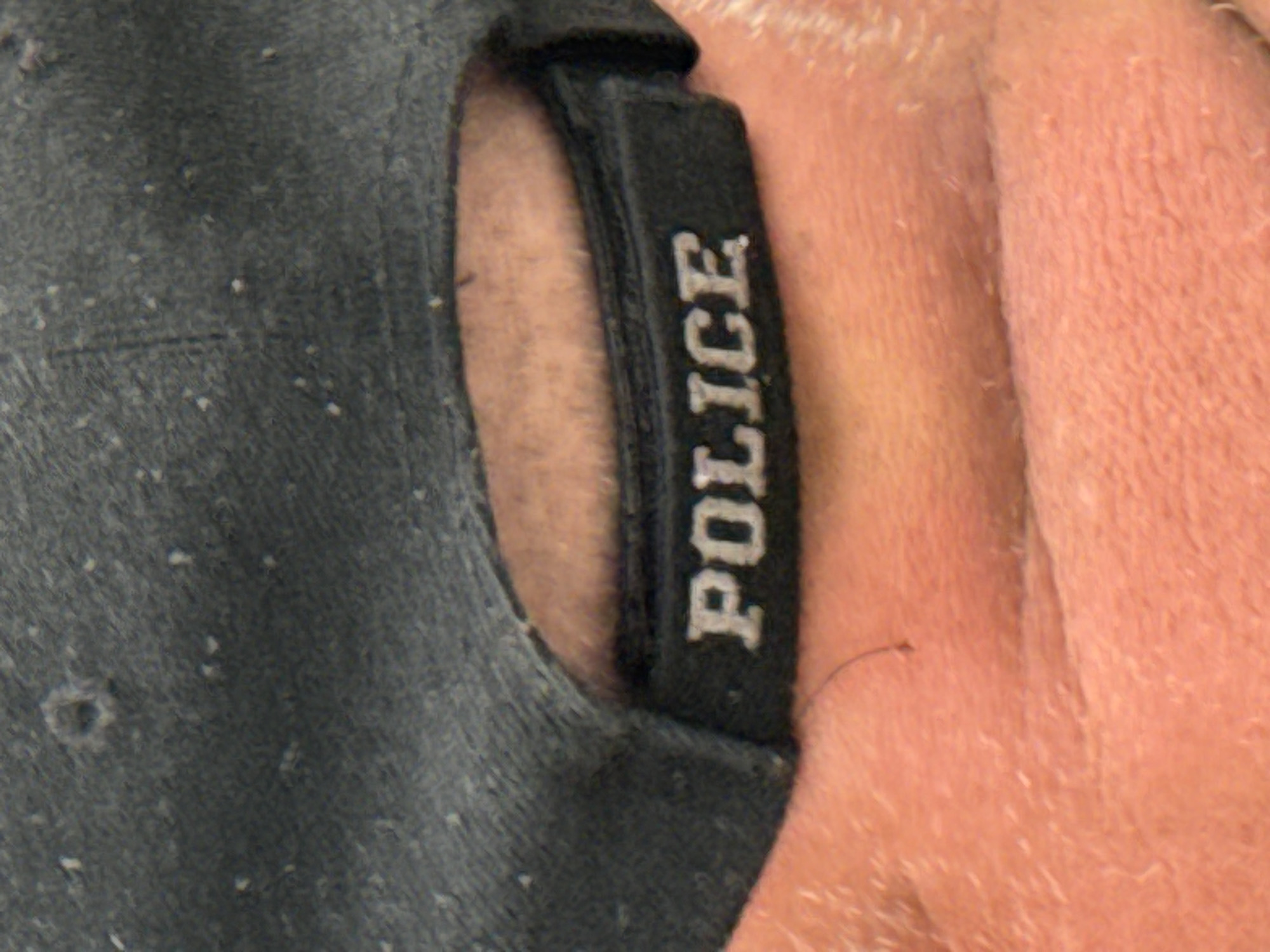 The image is a close-up, medium shot of the back of a dark grey baseball cap with the word "POLICE" embroidered in white, block letters on a black band. The cap is positioned on a person's head, and the brim of the cap is visible, along with a portion of the person's orange-toned skin, likely their neck or upper back, which appears to be sun-damaged or dry. The background is blurred, but the overall impression is of an outdoor setting. The exact location in Bellheim, Germany, is not discernible from the image alone.