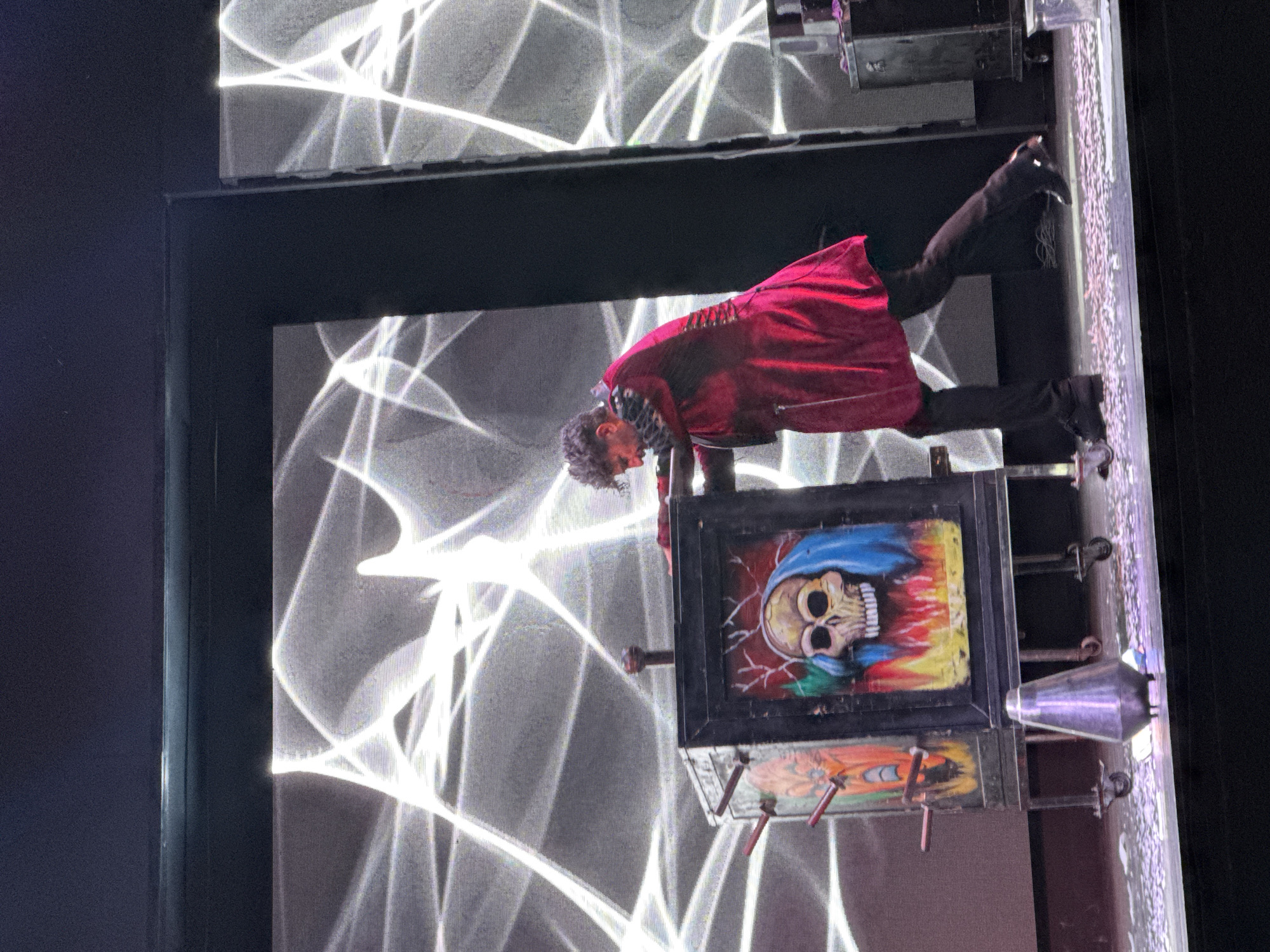 In Manavgat, Turkey, on a stage with a large screen displaying a white, web-like pattern, a magician performs a trick. He is wearing a striking red coat with fur trim on the collar and a corset-like detail on the back. The magician is interacting with a prop box that is decorated with images of a skull and a fiery background, along with another, less distinct image on the left side. The box is on wheels, suggesting it's part of the illusion. In front of the box, a metallic cone-shaped object rests on the stage, which is covered in confetti. The magician's body language indicates he is actively engaged in the performance, pushing the box.