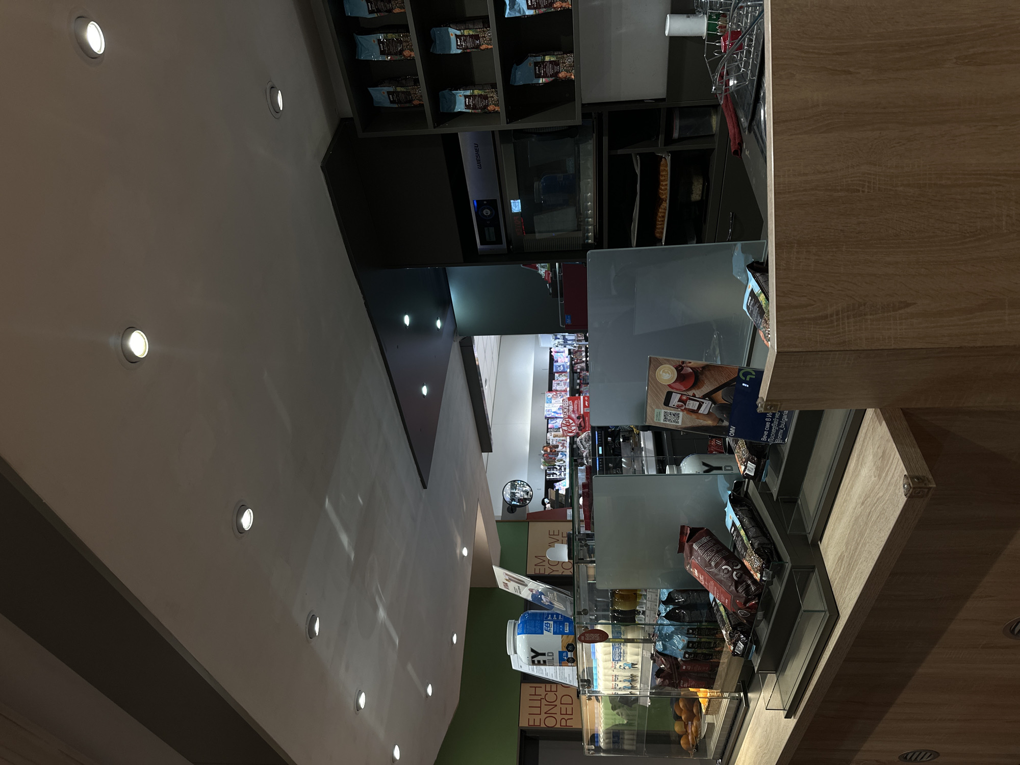 The image depicts the interior of what appears to be a café or convenience store, possibly a part of a gas station, located somewhere in Stara Zagora, Bulgaria. The scene is focused on a sales counter with a glass display case filled with various products, including snacks, protein powders, and packaged foods. A shelving unit behind the counter contains more merchandise. The brand "WIESHEU" is visible on what seems to be an oven or kitchen appliance. Above the counter, a menu board or promotional sign is displayed, featuring the words "OMV" and the phrase "Вече сме в Ин".
--- OCR End ---