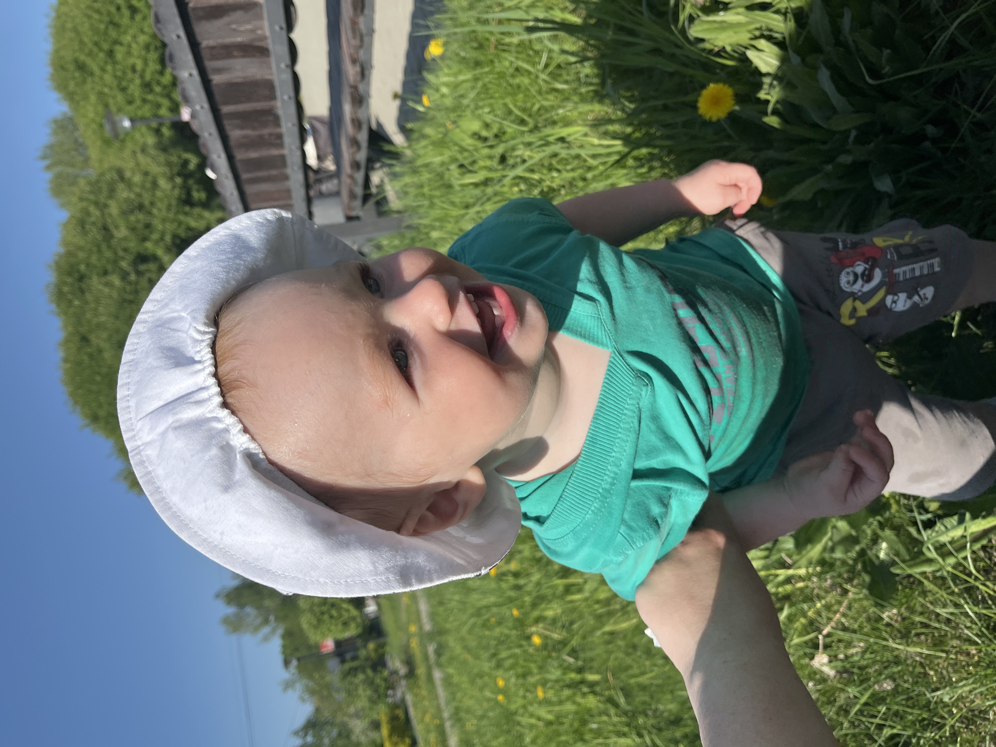 A baby wearing a white sun hat and a green t-shirt is being held in someone's arm outdoors on a sunny day. The baby is smiling and appears to be enjoying the moment. The background features green grass, some dandelions, trees, and part of a structure with a dark roof. The location is potentially Chaikovsky, Russia.