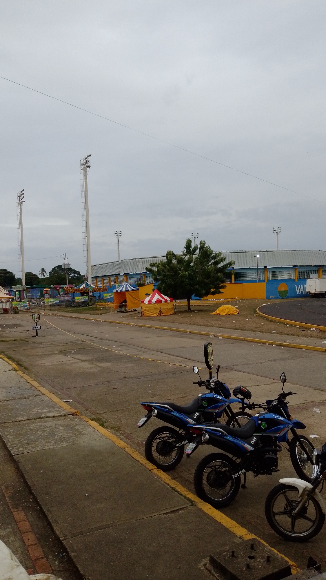 The image shows a street scene in Tinaquillo, Venezuela. In the foreground, three police motorcycles are parked along the curb. Behind the motorcycles is a paved area littered with debris, leading to what appears to be a stadium or event venue. Several tents and structures are visible within the venue area. Light towers stand tall near the stadium. The sky is overcast.