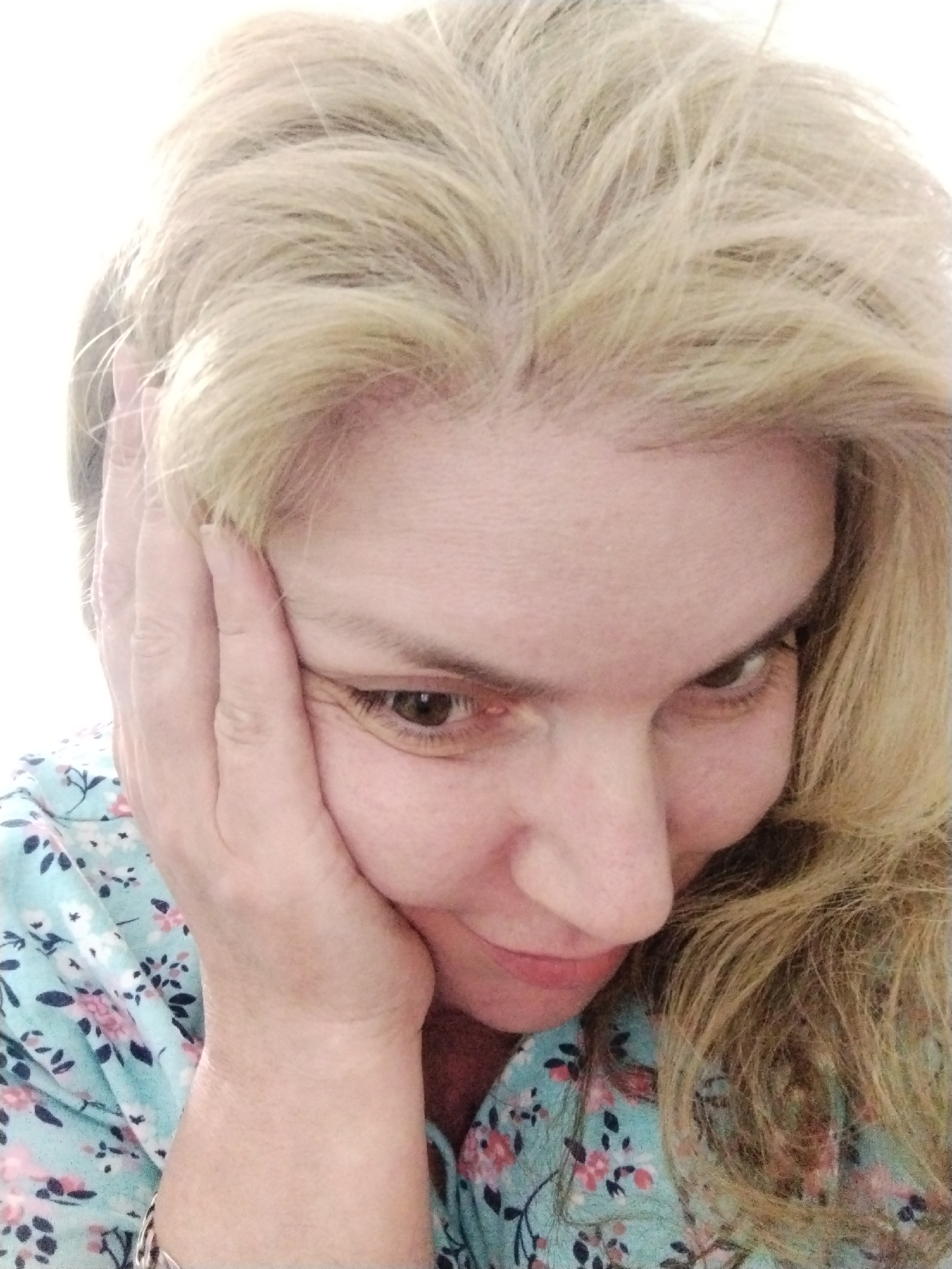 A close-up, overhead shot shows a woman with light blonde hair resting her face in her left hand. She is wearing a light blue shirt with a floral pattern. The background is a plain white. The image is a selfie taken in Al Mas'oudi, United Arab Emirates.