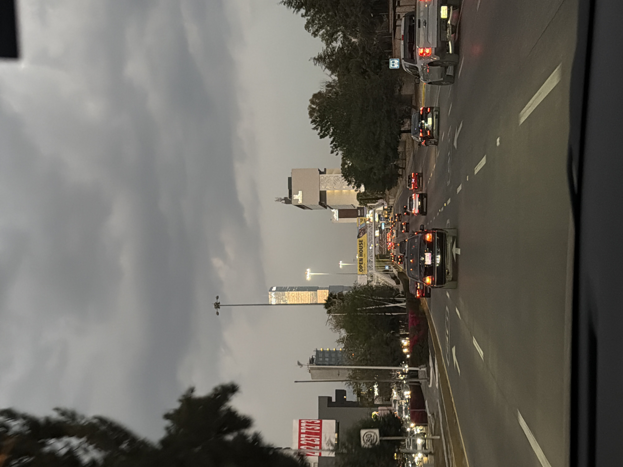 The image displays a multi-lane urban thoroughfare in San Bernardino Tlaxcalancingo, Mexico, viewed from inside a vehicle at dusk or under heavily overcast conditions. The sky is a uniform gray. No people are visible.
The roadway features multiple lanes separated by white dashed and solid lines, with directional arrows painted on the asphalt indicating forward movement. Numerous vehicles are present in both directions, with their headlights and taillights illuminated, suggesting active traffic flow. A dark-colored sedan occupies the center foreground lane, while a silver pickup truck is visible in the rightmost lane further back.
On the left side of the image, a partial view of a dark tree obscures the immediate foreground. Beyond it, a white billboard with red text displaying "2 237 7373" is prominent. Below the billboard, a circular speed limit sign is visible. Tall streetlights with multiple lamp fixtures line the road.
The mid-distance and background feature several modern, multi-story buildings. A tall, glass-clad skyscraper bearing the "BBVA" logo near its summit reflects ambient light. To its right, another contemporary building is visible. An overhead structure, possibly a pedestrian bridge or an overpass, spans the road, displaying a yellow sign with "OPEN HOUSE" in black lettering. Dense trees and bushes line both sides of the road, contributing to the urban landscape. The bottom edge of the image reveals the upper portion of a vehicle's dashboard.