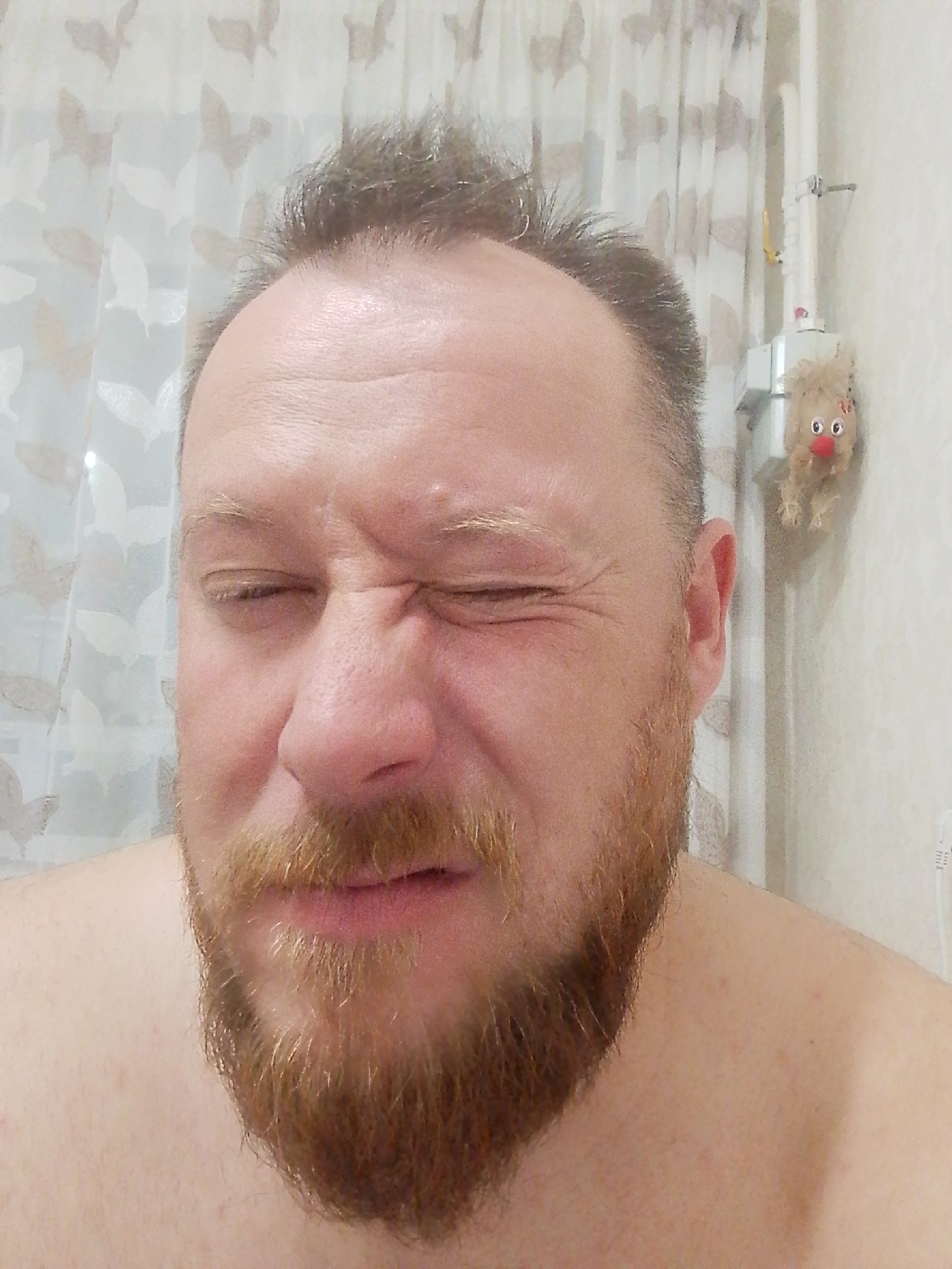 A close-up, full-frontal image shows a middle-aged, fair-skinned man with a reddish-brown beard and thinning hair squinting tightly with his eyes closed. He is photographed from the chest up, bare-chested. Behind him is a patterned white and beige curtain. A small stuffed animal resembling a reindeer hangs on the wall near his right ear. The location, Фоки, Russia, is not discernible from this image.