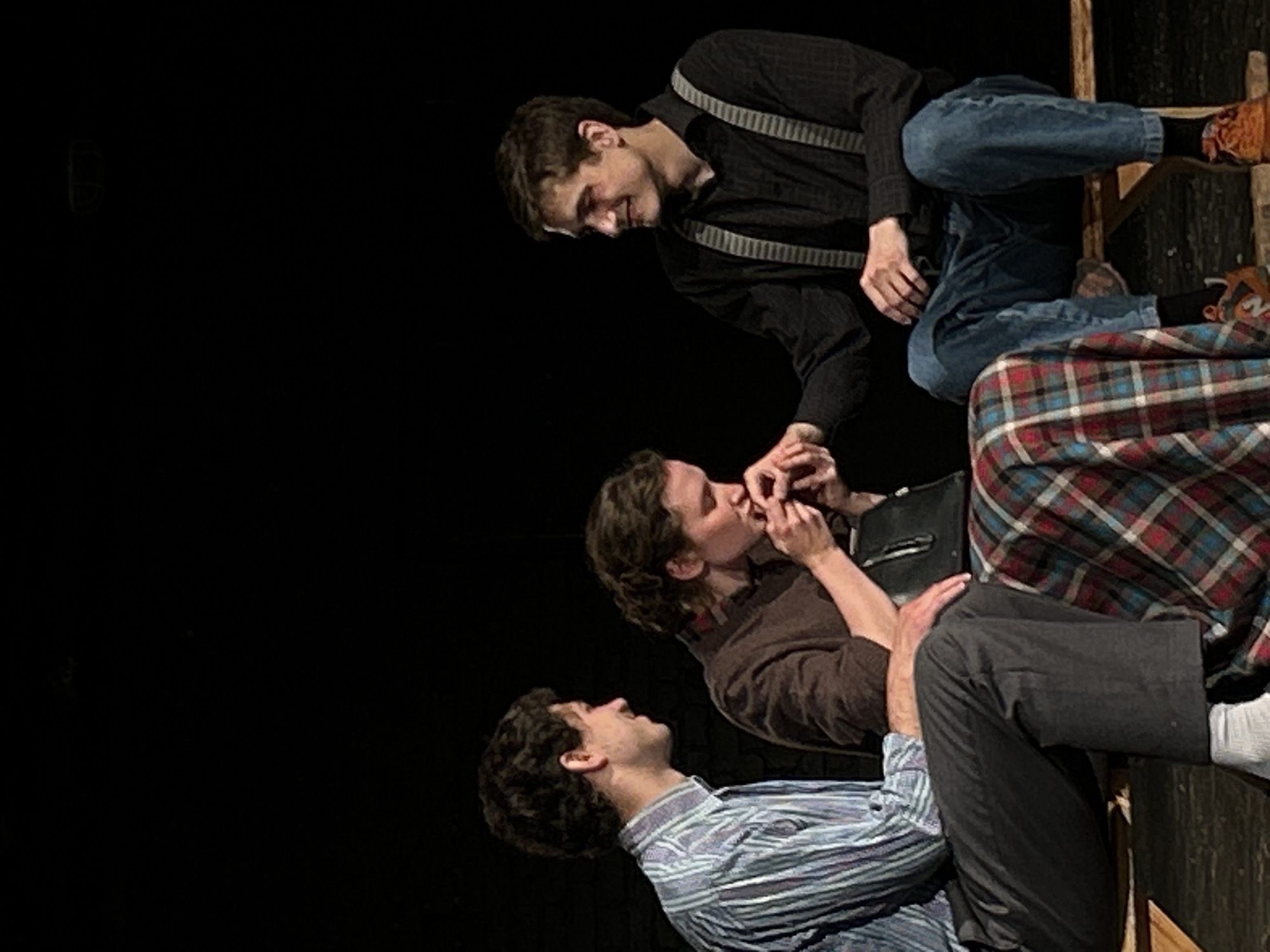 Three individuals are seated on a wooden bench against a black background. On the left, a man with curly brown hair wears a light blue and white striped long-sleeved shirt and dark gray trousers, with white socks. In the center, a woman with brown curly hair wears a brown sweater over a plaid skirt and holds a black purse. She is kissing the hand of the man on the right. The man on the right has brown hair and wears a black collared shirt with suspenders and blue jeans, along with orange sneakers. The scene appears to be a theatrical performance or a posed photograph.