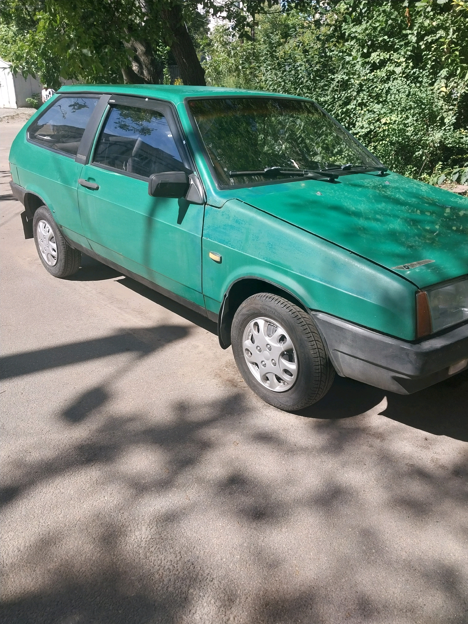 An eye-level, medium shot captures a weathered, green, three-door hatchback car parked on an asphalt surface in a city setting. The car, likely a Soviet-era model, exhibits signs of age with faded paint and a visible matte finish on the hood. Sunlight casts dappled shadows from overhead foliage onto the car and the road, suggesting a bright, daytime scene. The background is dominated by dense green foliage and a large tree with a textured trunk. In the upper left corner, a white building with a sloped roof is partially visible, and a small section of what appears to be a street sign is present. The overall environment suggests a quiet, residential street or parking area within a city like Dnipro, Ukraine.