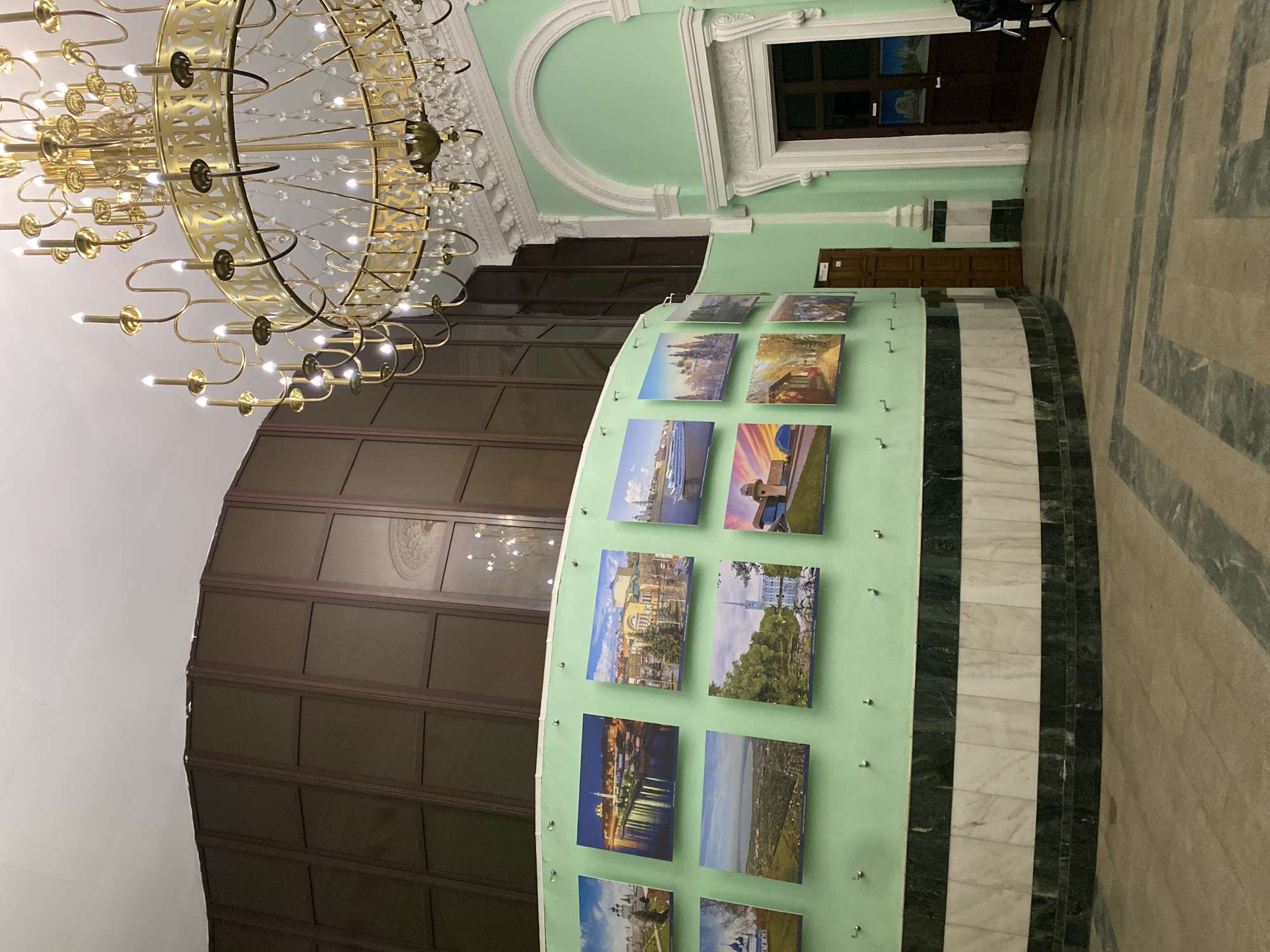 An interior view of a grand hall or public space is captured from a low angle, dominated by an ornate, multi-tiered gold chandelier in the upper left, featuring numerous bulbs and crystal accents. The ceiling is white with some ornate white molding, extending to a pastel green wall on the right.
A prominent, curved, light green partition or display wall runs through the mid-ground, serving as an exhibition space. It showcases multiple square photographs or postcards arranged in rows. These images depict various landscapes and cityscapes, including a boat on a river, architectural landmarks, urban scenes, and a city reflecting lights on water at night. The base of this curved wall features elegant stripes of white and dark green or black marble.
Further right, the light green wall continues with elaborate white classical architrave detailing around a window or recessed opening, and a dark wooden door is visible below. The floor is tiled with light-colored marble or stone, exhibiting a geometric pattern. To the left of the green partition, a large, dark brown paneled or slatted structure rises, possibly a wall or room divider, reflecting parts of the room. No people are visible, and there is no discernible text on the images or in the environment. The artificial lighting from the chandelier suggests an indoor setting, possibly a gallery, hotel lobby, or public building.