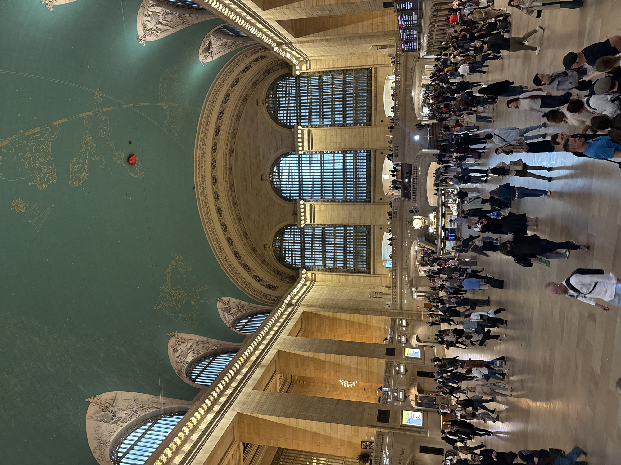 The image shows the interior of Grand Central Terminal in New York City. The ornate ceiling, painted with constellations, dominates the upper portion of the image. Below, large arched windows let in natural light. The main concourse is filled with people, some appearing to walk in various directions, and some appear to be standing. Ticket kiosks and information boards are visible along the sides. The overall environment suggests a busy transportation hub.