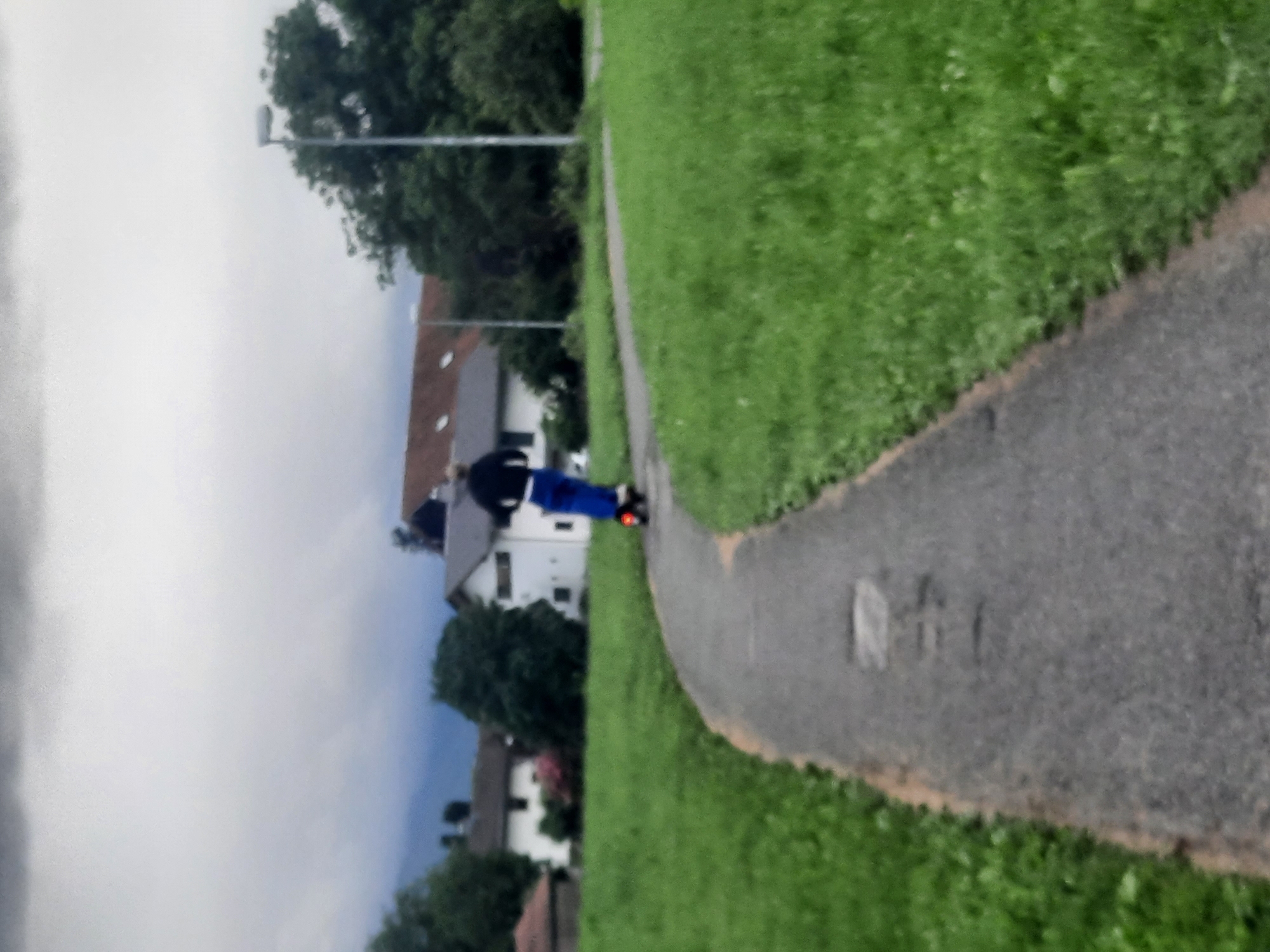 A person is riding an electric scooter on a paved path that curves through a grassy area. The person is wearing a dark jacket and blue pants, and the scooter has a red light on the back. In the background, there are several houses with dark roofs, trees, and streetlights. The sky is overcast and appears cloudy. The overall scene depicts an outdoor recreational setting.