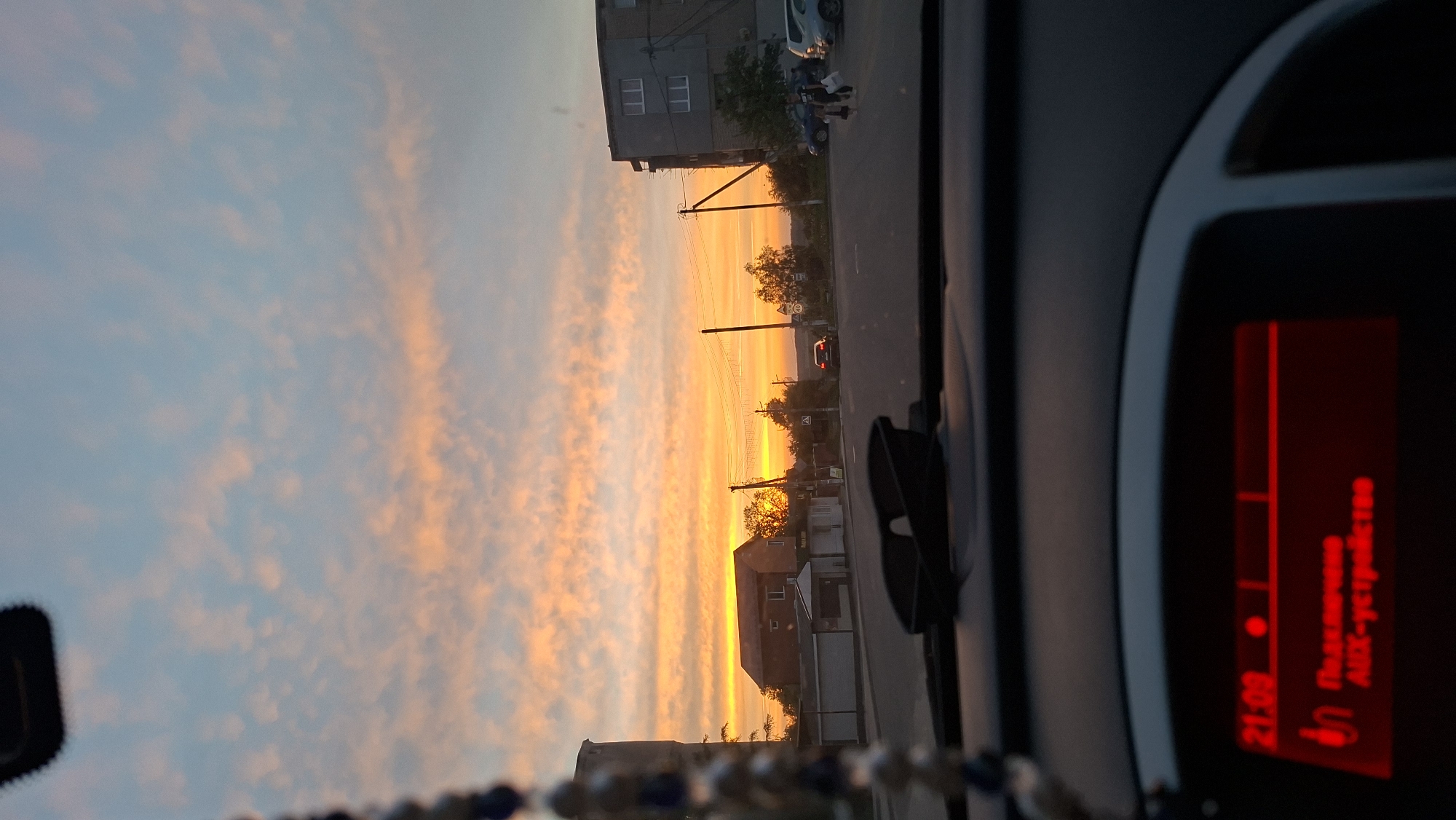 The image shows a view from inside a car, likely the dashboard, with a sunset visible through the windshield. The location is a street in a town or city with buildings, utility poles, and a road. The sky is filled with clouds, with a strong orange and yellow glow from the setting sun. The dashboard features a digital display showing "21:08" and text in a Cyrillic alphabet. There is a phone mount on the dashboard. Some blurred beads hang from the rearview mirror. Pedestrians are crossing the street in the distance.