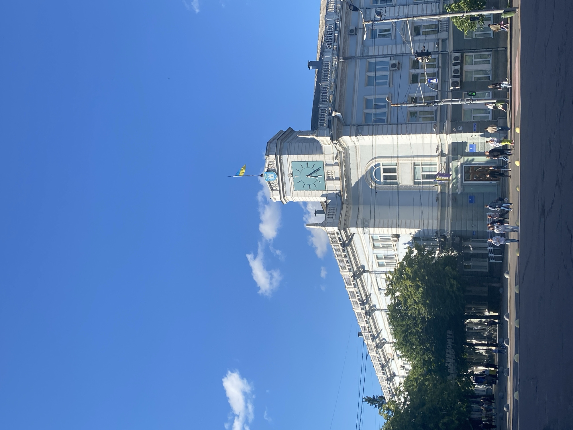 The image shows a street scene in front of a light blue, multi-story building with a prominent clock tower. The building has a long facade with numerous windows and a decorative balustrade along the roofline. A Ukrainian flag flies from the top of the clock tower, which features a large, light blue clock face. The sky is clear blue with a few white clouds. In front of the building, there are trees lining the sidewalk, and a street with cars and pedestrians. Several people are walking on the sidewalk and crossing the street. A traffic light is visible on a pole. The ground in the foreground is a dark asphalt surface with round, light-colored bollards. The overall impression is a bright, sunny day in an urban setting.