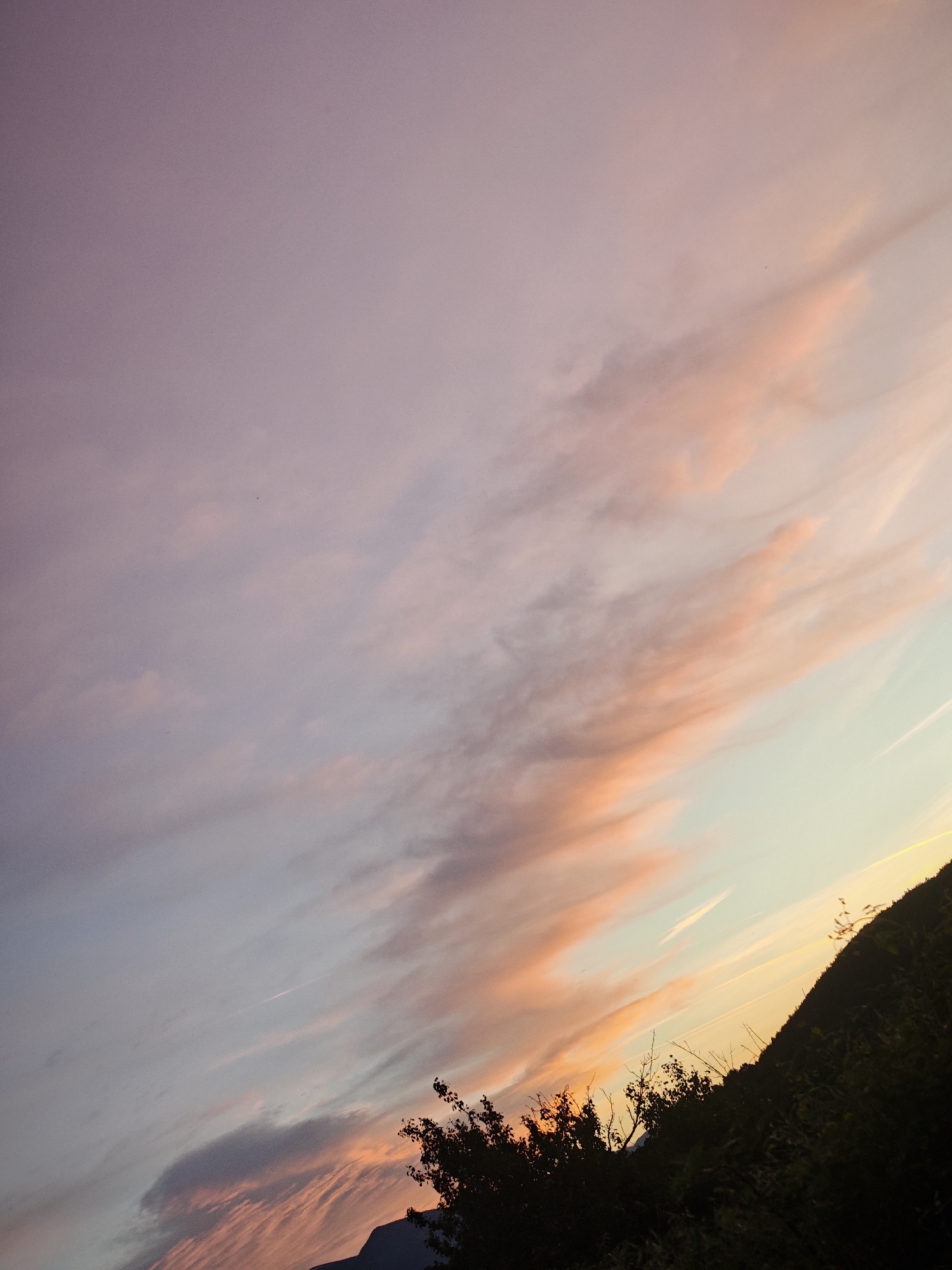 The image depicts a sky at dusk in Dabravite, Bulgaria. The sky is a blend of pink, peach, and light blue hues, with clouds stretched across the frame. A dark silhouette of a hill or mountain is visible in the lower right corner. The outlines of trees are also visible along the base of the hill and lower left.