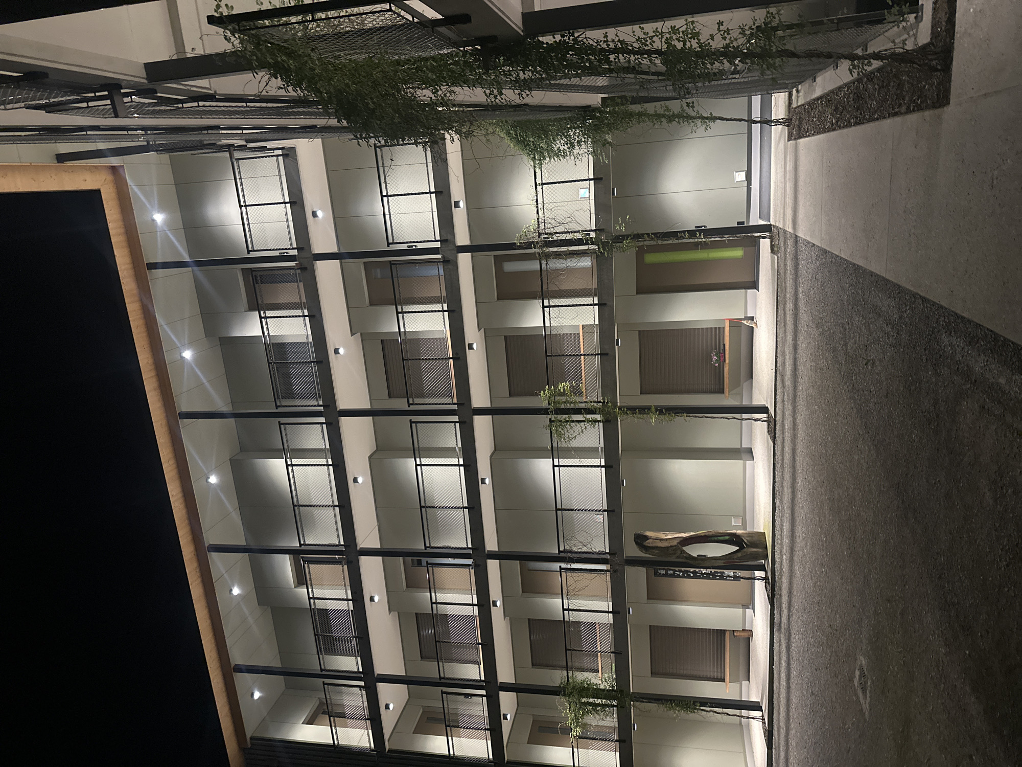 The image shows a multi-story building facade at night, featuring a grid-like structure with balconies or open spaces. The building is pale in color, with dark window coverings. There are plants or vines hanging from the upper levels. A pathway runs alongside the building, with a planter area on the right. A large, hollowed-out log structure serves as a bench or decorative feature near one of the entrances. The location and city of Fehraltorf, Switzerland, cannot be confirmed, as there is no additional context to corroborate this information.