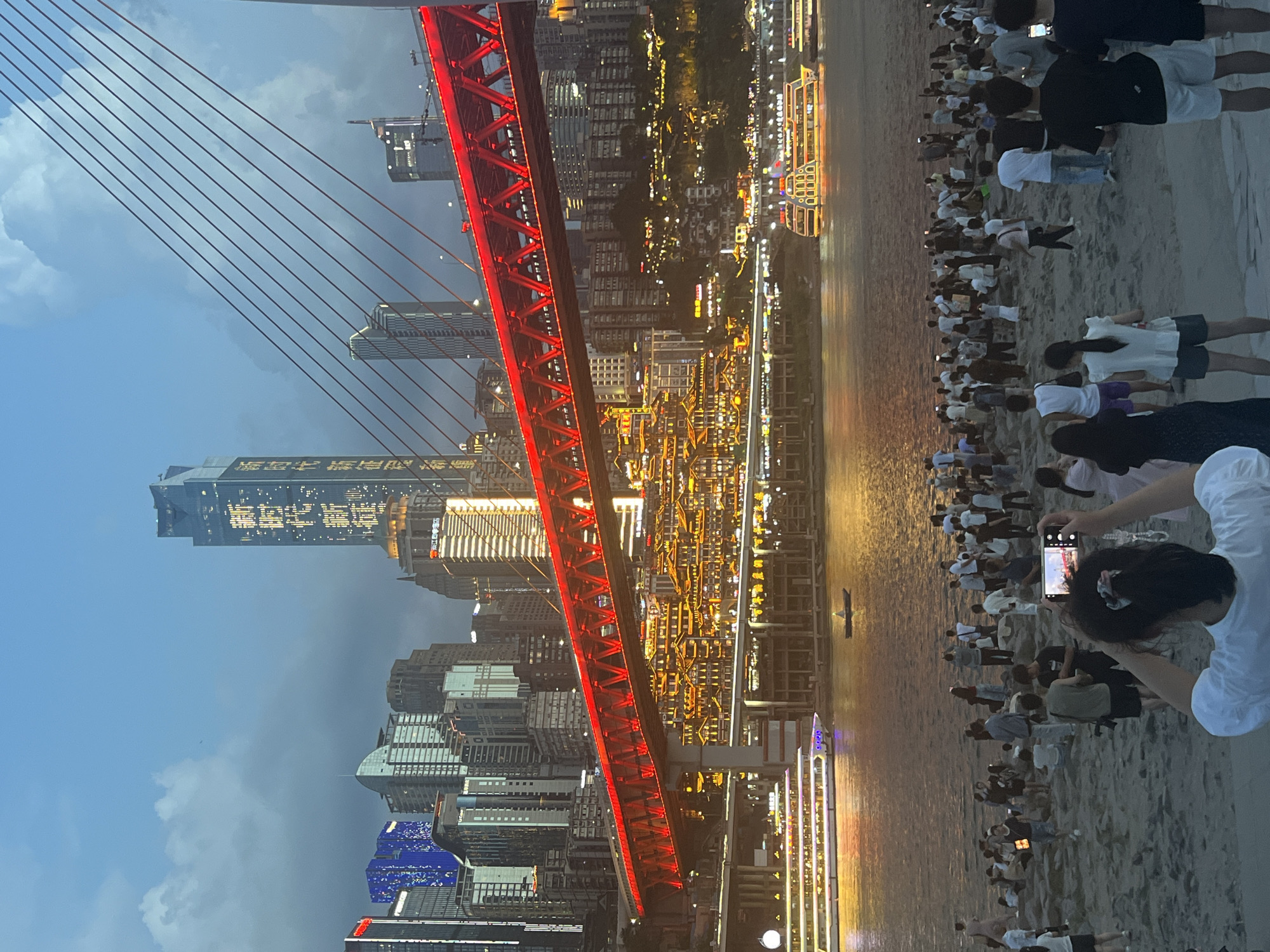 The image depicts a wide shot of the Jiangbei Cheng Street, China, waterfront at dusk. A prominent red cable-stayed bridge spans across the river, illuminated with bright lights. Behind the bridge, a dense cityscape of modern skyscrapers, some featuring glowing Chinese characters, rises against a twilight sky. Traditional-looking buildings with numerous illuminated windows are clustered along the riverbanks below the skyscrapers.
In the foreground, a sandy embankment is populated by a large group of people. Many are standing, some are sitting, and a few are walking. Most individuals are dressed casually in summer attire. A woman in the immediate foreground is holding up a smartphone, appearing to take a photograph of the bridge and cityscape.
On the river, several boats are visible. A large, multi-deck passenger boat is docked or slowly moving near the embankment, its windows and exterior brightly lit. Further out on the water, a smaller, traditional-style boat with a single occupant is also present. The river's surface reflects the lights of the city and the sky. The overall atmosphere suggests a popular tourist or recreational area in the early evening.