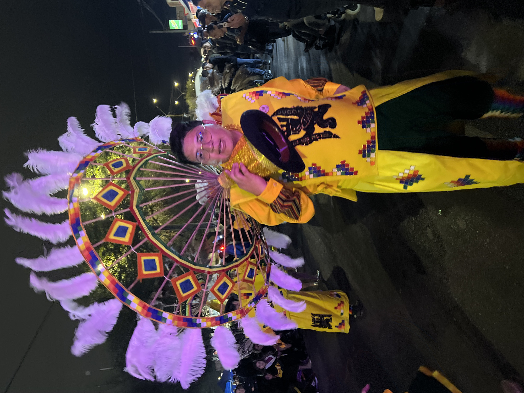 A person in a colorful yellow costume with a large feathered headdress is posing for a photo at night in Arica, Chile. The headdress features a circular frame adorned with colorful diamond-shaped patterns and large white feathers. The costume includes a long yellow tunic with intricate geometric patterns and a wide, dark hat held in one hand. The person is wearing glasses and looking at the camera. In the background, other people in similar attire and a crowd are visible, suggesting a parade or festival. The scene is lit by streetlights, and a portion of a building with a green sign is visible on the right. The ground appears to be asphalt.