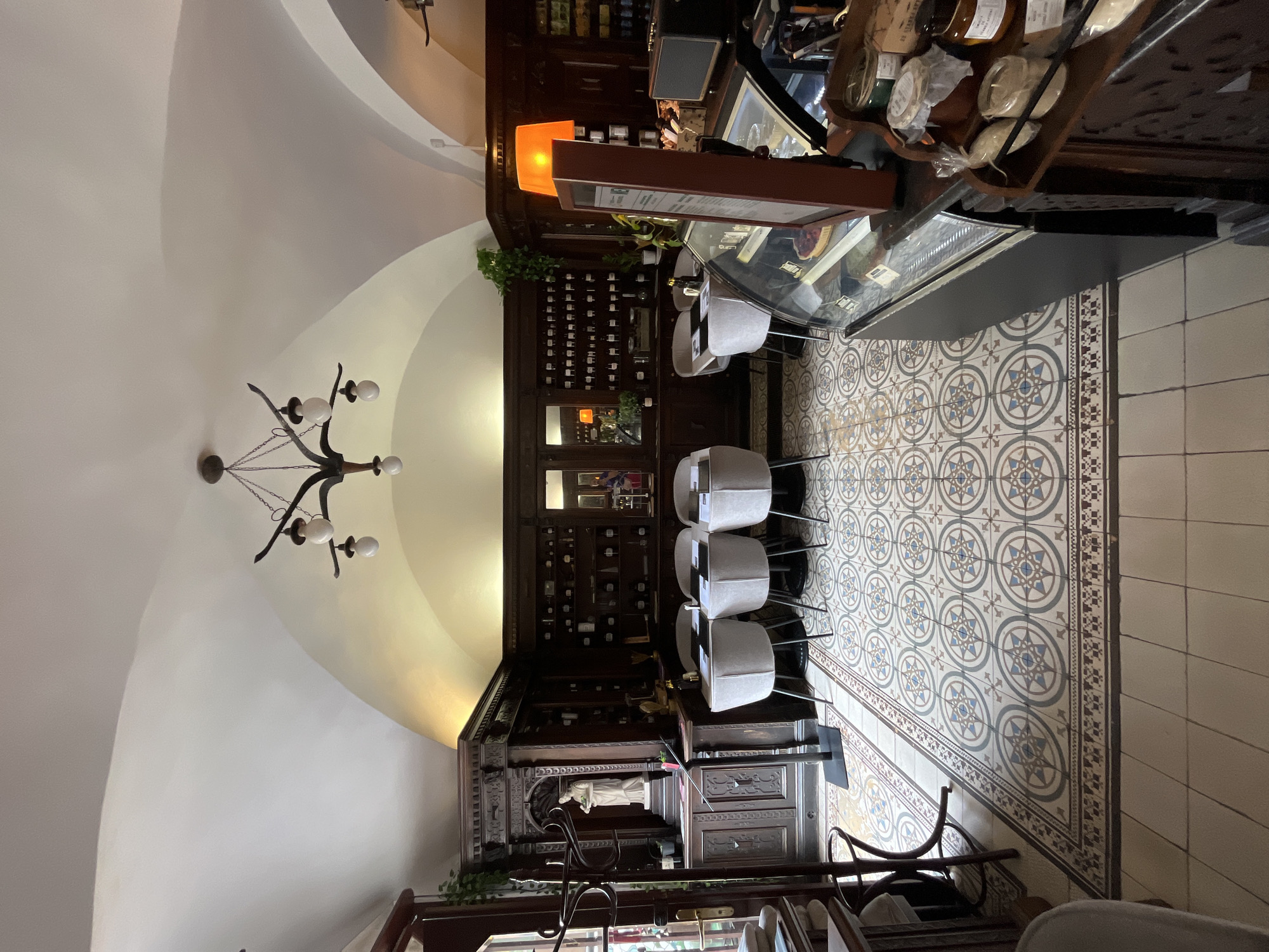 An interior shot of a cafe or restaurant shows a dimly lit room with vaulted ceilings and a patterned tile floor. The room is furnished with several tables and chairs, a display case with food items, and shelves stocked with bottles. The decor suggests an older, possibly European, establishment. The lighting is warm, with an orange lamp adding to the ambient glow. The scene is calm and quiet, with no people visible.