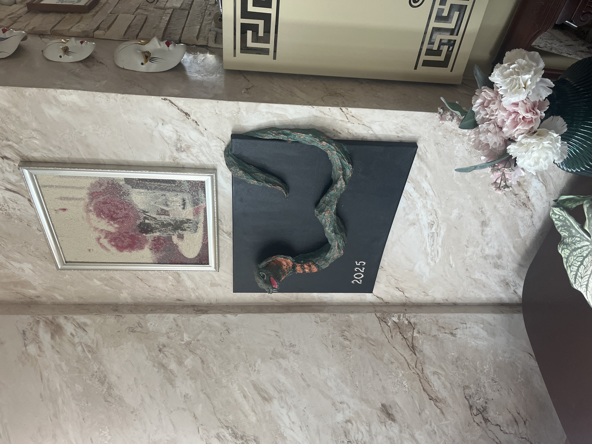 The image shows an interior scene with decorative elements on a wall. A framed artwork, possibly a diamond painting of flowers in a vase, hangs on the upper portion of the wall. Below it, a dark rectangular plaque features a three-dimensional sculpture of a green snake coiled across it. The number "2025" is inscribed in white at the bottom left of the plaque. To the right of the plaque, a series of white decorative masks with red lips and gold accents are mounted on the wall. Further to the right, a portion of a light-colored cabinet with a Greek key design is visible. In the foreground, a dark piece of furniture, possibly a table, is partially visible, along with a vase of artificial flowers in shades of pink and white. The wall has a textured finish resembling marble. There are no people present in the image. The location is stated to be Vladimirovka, Russia.
