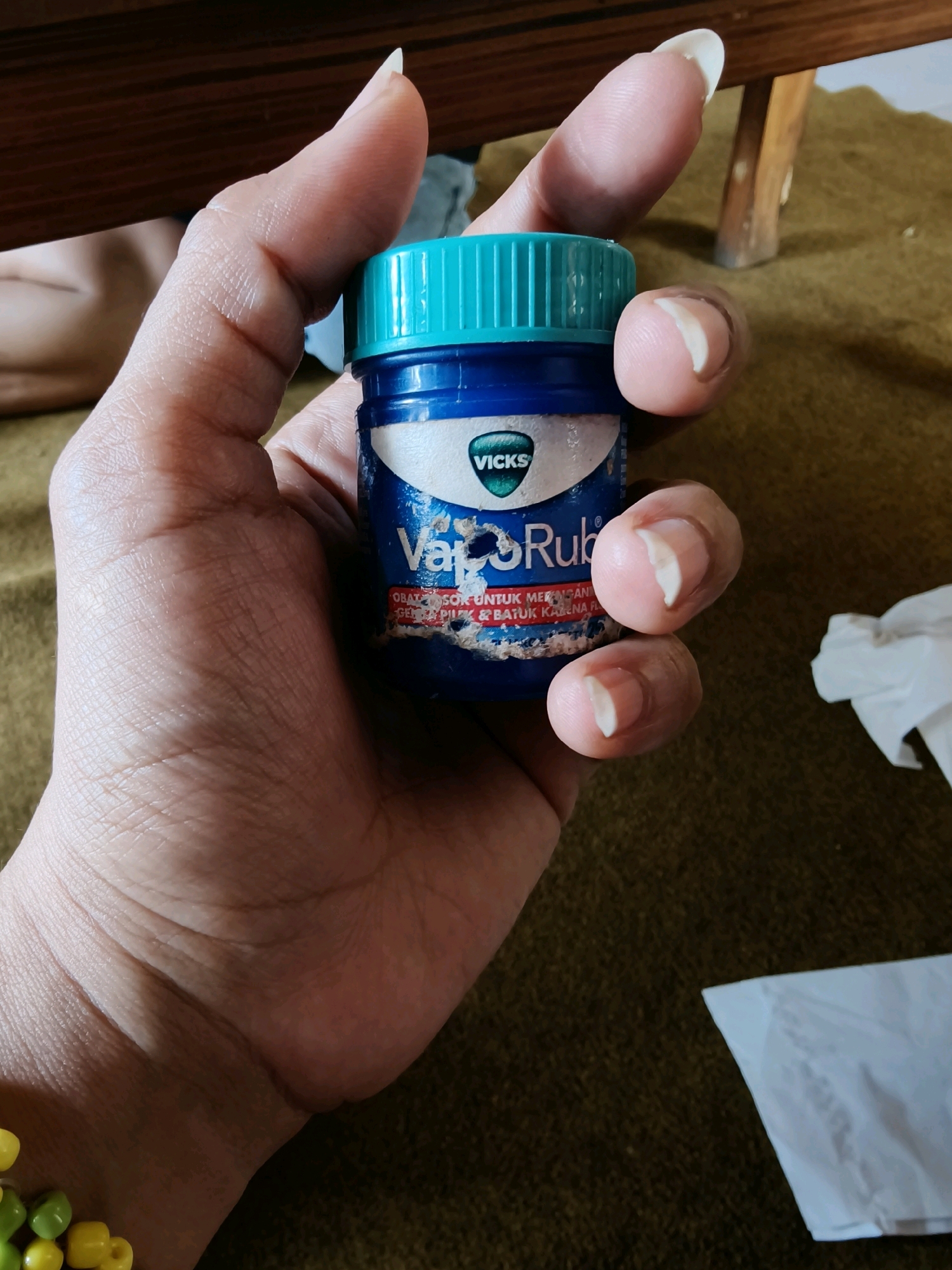 In Ploso, Indonesia, a hand holds a small blue Vicks VapoRub jar. The jar has a teal-colored screw-top lid and a label that reads "Vicks" above "VapoRub" with some text below in Indonesian. The hand's fingers, with light-colored nail polish, grip the jar, obscuring part of the label. The background consists of a textured brown surface, possibly a carpet or rug, with a wooden furniture leg visible in the upper right corner. A crumpled piece of white tissue paper lies on the surface to the right of the hand.