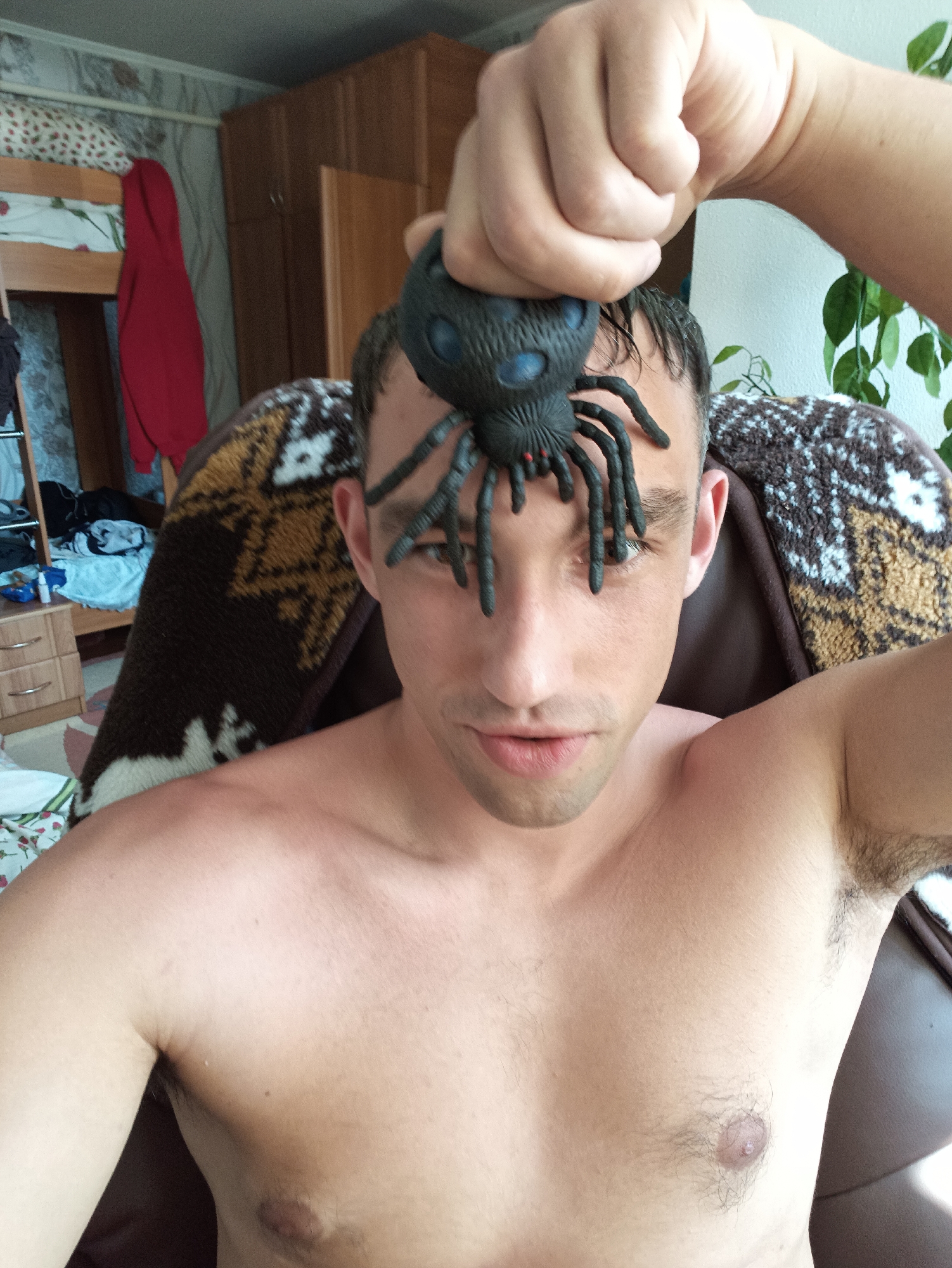 The image depicts a man, shirtless, holding a black toy spider above his forehead. He is seated in a chair with a patterned blanket draped over it. The background suggests an indoor setting, possibly a bedroom, with a bunk bed, wardrobe, and drawers visible. A red garment hangs from the bunk bed. Plants are visible in the upper right corner. The man's skin tone is fair, and he has short, dark hair. His expression is neutral. The image quality is moderate, with some graininess. This scene is set in Berezne, Ukraine.