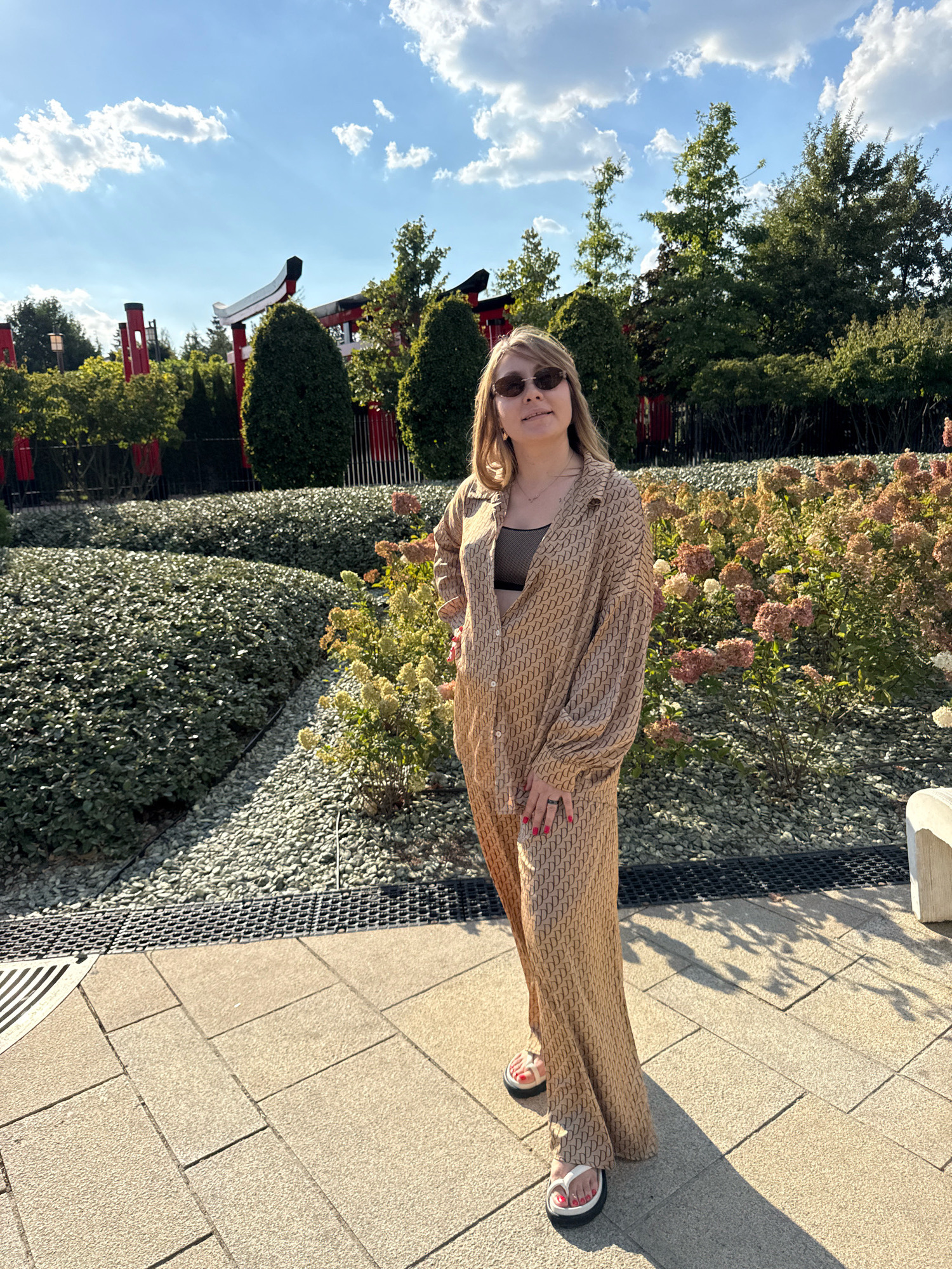 A young woman with blonde hair and sunglasses stands in an outdoor setting. She is wearing a tan, patterned pantsuit and a black top. The background features manicured bushes, gravel pathways, trees, and structures resembling traditional Japanese torii gates. The scene appears to be a park or garden, possibly in Krasnodar, Russia, given the context. The sky is blue with scattered clouds. The woman is looking towards the camera and has her hands on her hips. The lighting suggests it is daytime with sunlight casting shadows.