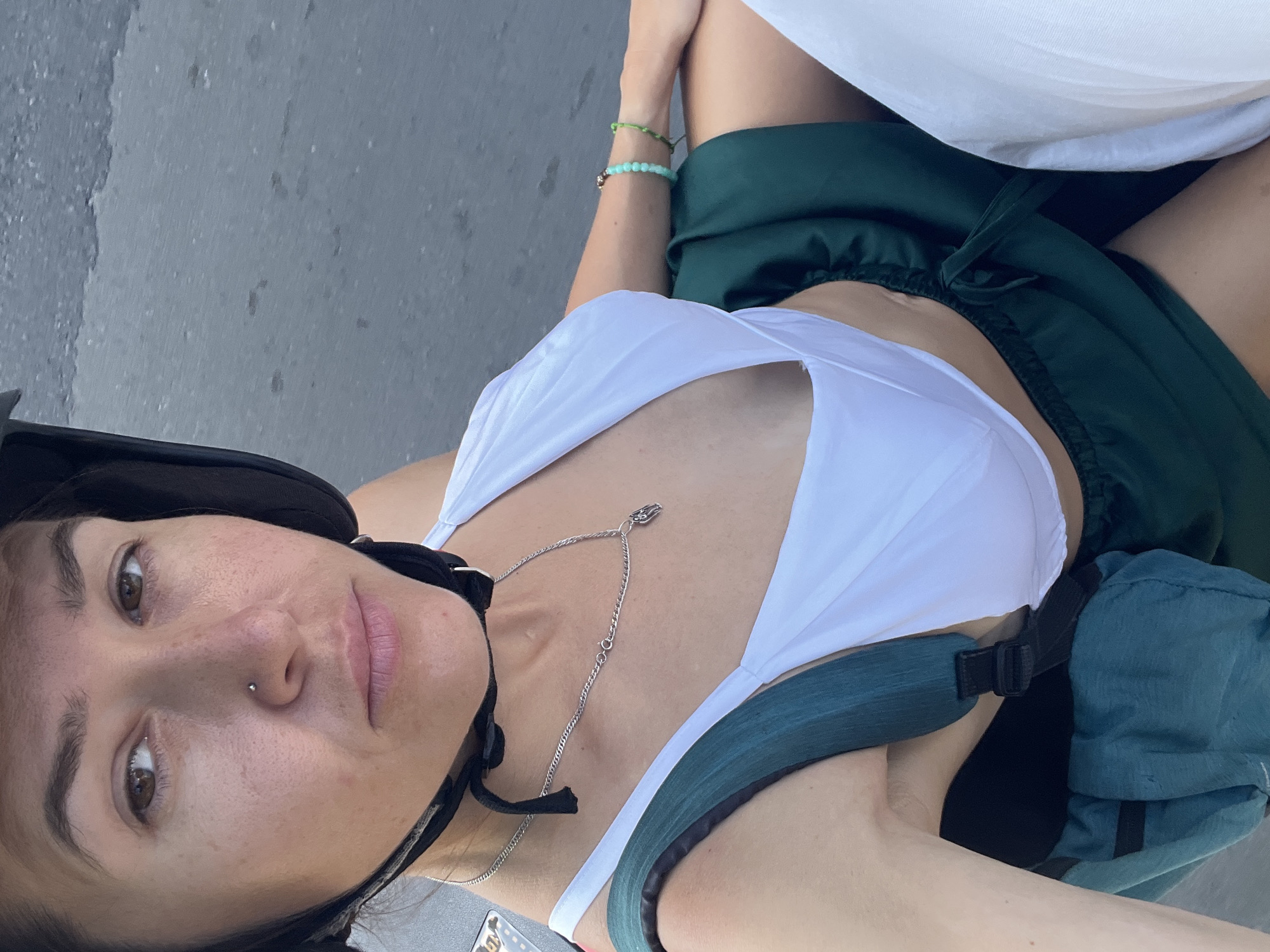 A woman wearing a white bikini top, a dark green skirt, and a black helmet is pictured from the chest up. She is wearing a silver necklace with a small charm. On her right wrist, she has two beaded bracelets. A dark green backpack is visible on her left side. The background appears to be a paved road, suggesting an outdoor urban environment.