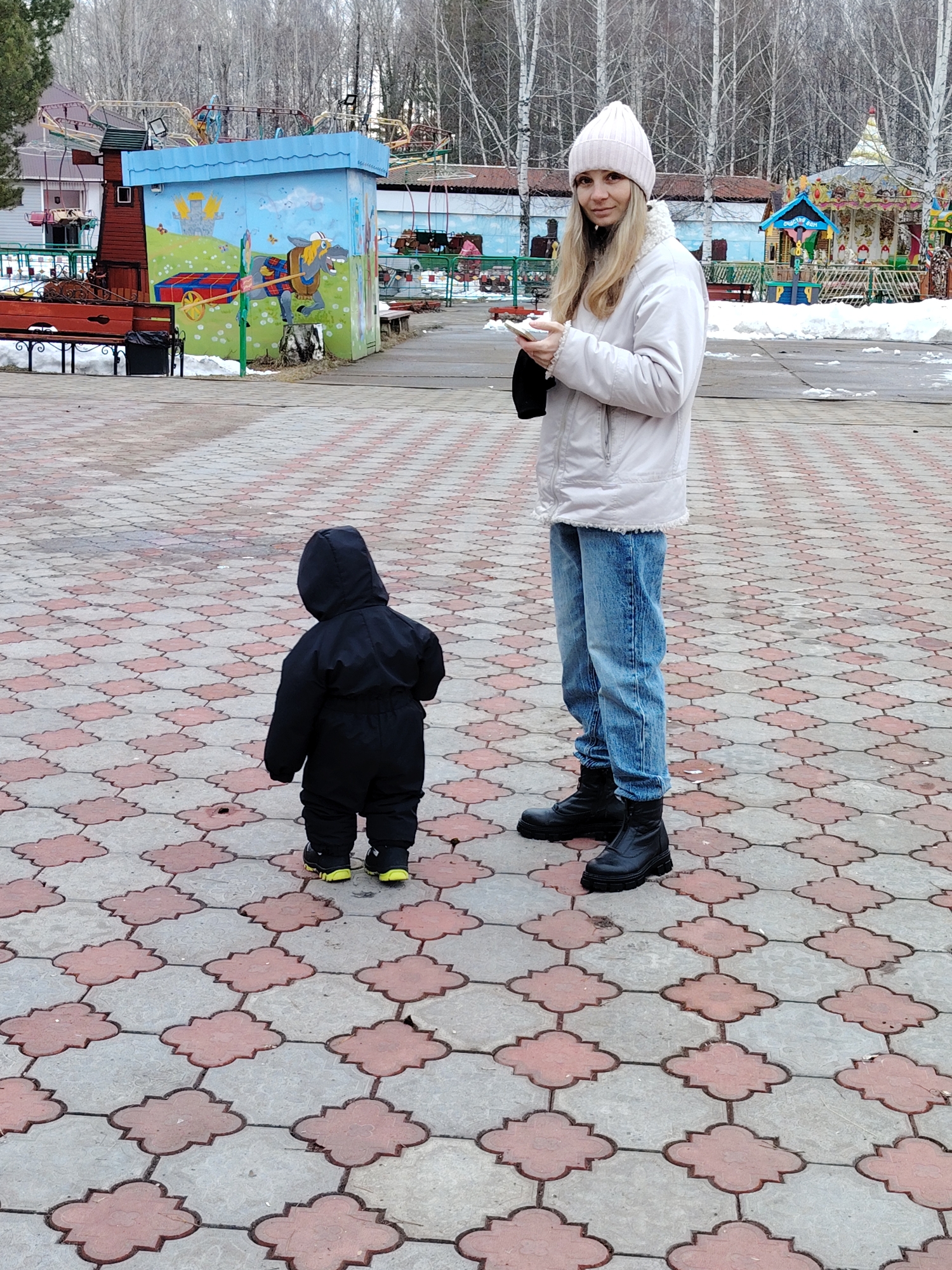 In the city of Chaikovsky, Russia, a woman and a young child are standing on a paved area with a distinct geometric pattern of grey and reddish-brown interlocking shapes. The woman is wearing a pale pink knit hat, a light-colored jacket, blue jeans, and black boots. She is holding a smartphone in one hand and a dark-colored object, possibly gloves, in the other. The child, facing away from the camera, is dressed in a full-body black suit with a hood. The child's shoes have a bright yellow trim.
In the background, there are various park structures including a small building adorned with colorful murals and several amusement park rides. Bare trees are visible behind these structures, suggesting it is either late autumn or winter. A bench is located to the left side of the image and patches of snow can be seen near the park structures.