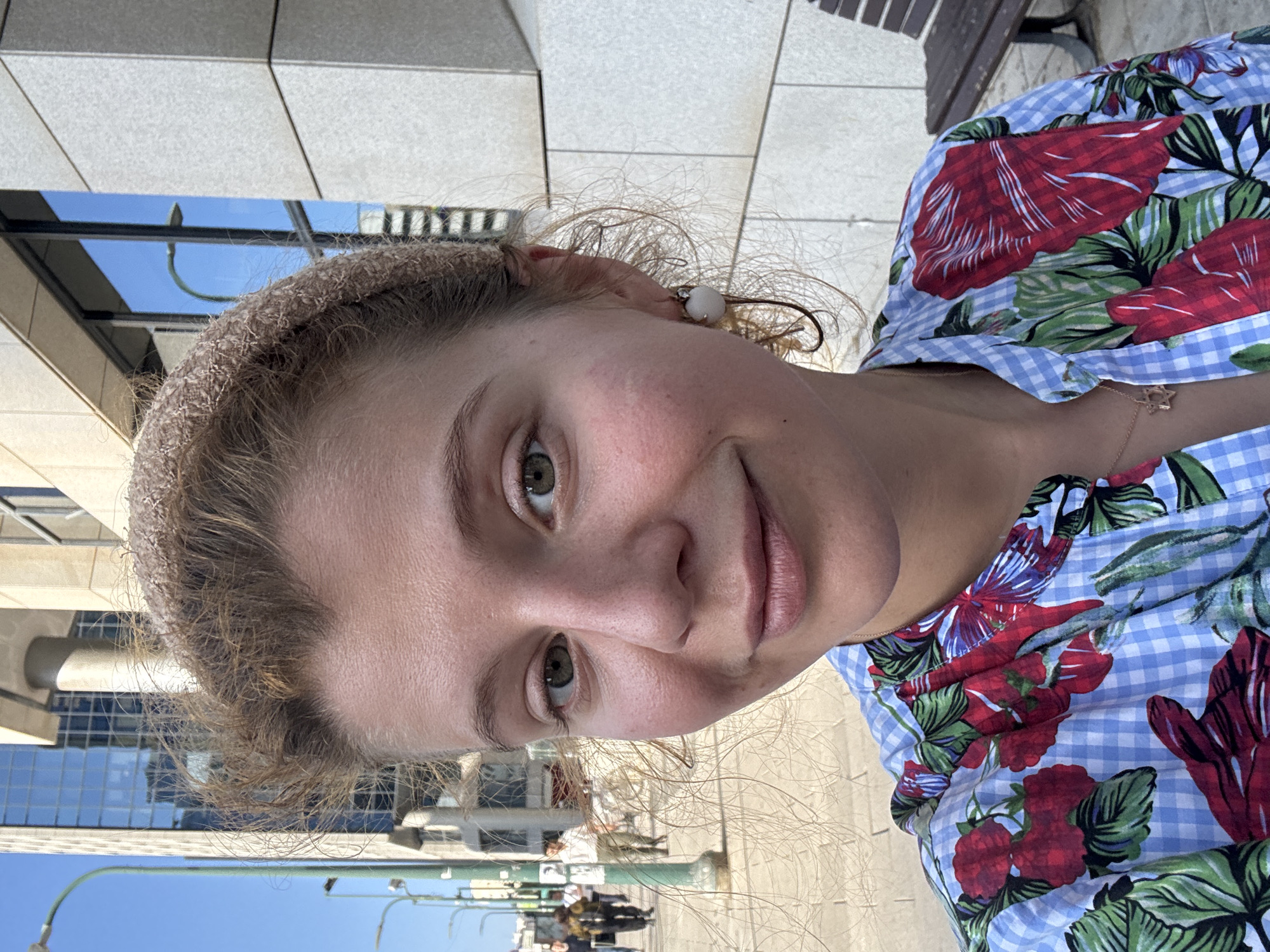 This is a close-up, sideways selfie of a young woman with light brown, curly hair. She is wearing a light brown, textured headband and a blue and white checkered shirt with a red and green floral print. A thin, gold necklace with a star pendant is visible on her neck. Her right ear is adorned with a pearl-like earring. She has light green eyes, light eyebrows, and her lips are slightly parted. The background shows a modern building with large windows reflecting a clear blue sky. There are also glimpses of a tiled walkway, a park bench, and some distant figures on a street. The lighting suggests it is daytime, likely sunny given the bright sky and reflections. The overall impression is casual and relaxed.