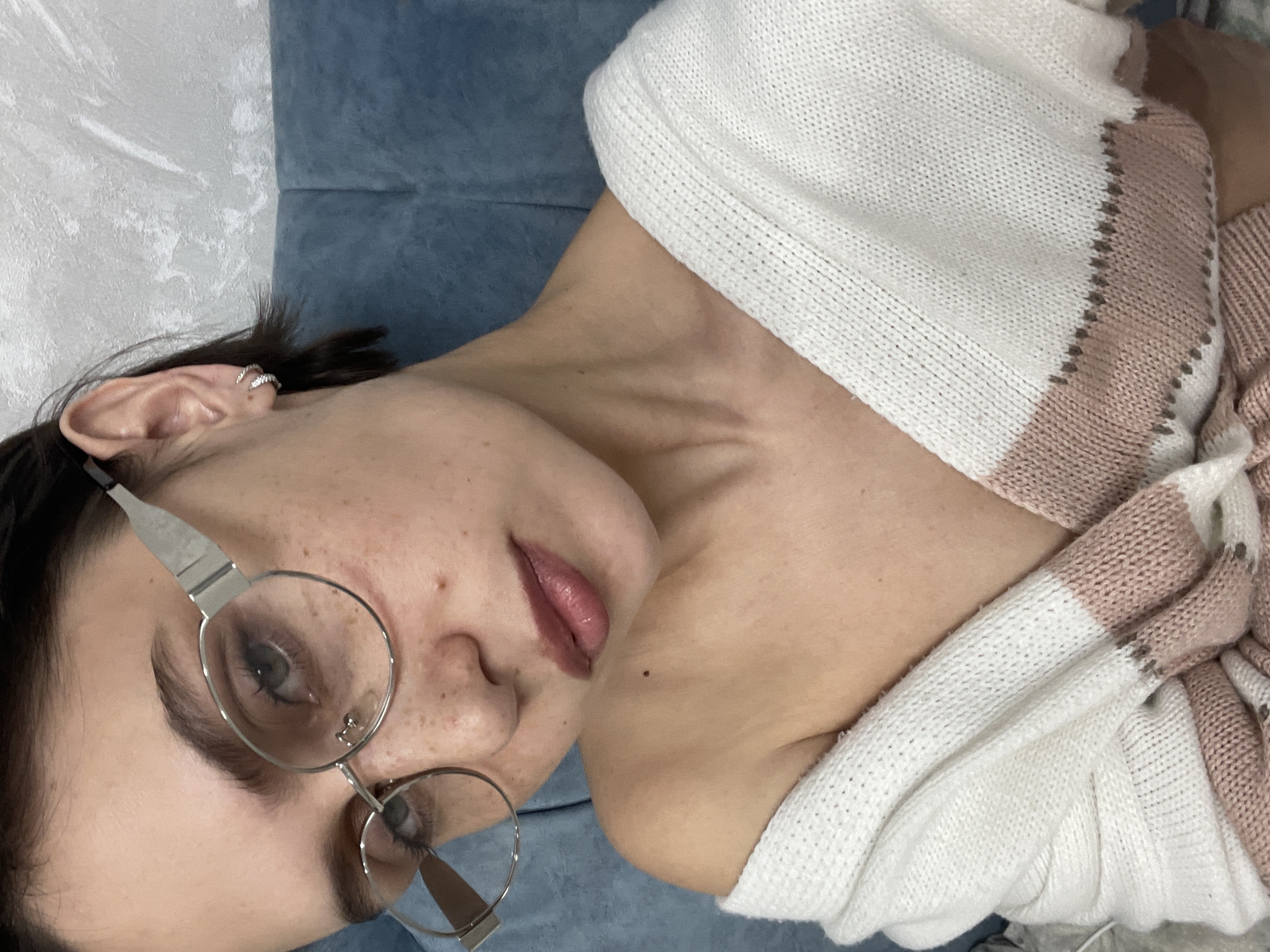 The image is a close-up selfie of a woman with fair skin, freckles, and dark hair. She is wearing round, silver-rimmed glasses. She has jewelry in her ear. She is wearing a white and pink striped sweater. The background is a solid light blue. There is no indication of the location being Izhevsk, Russia.