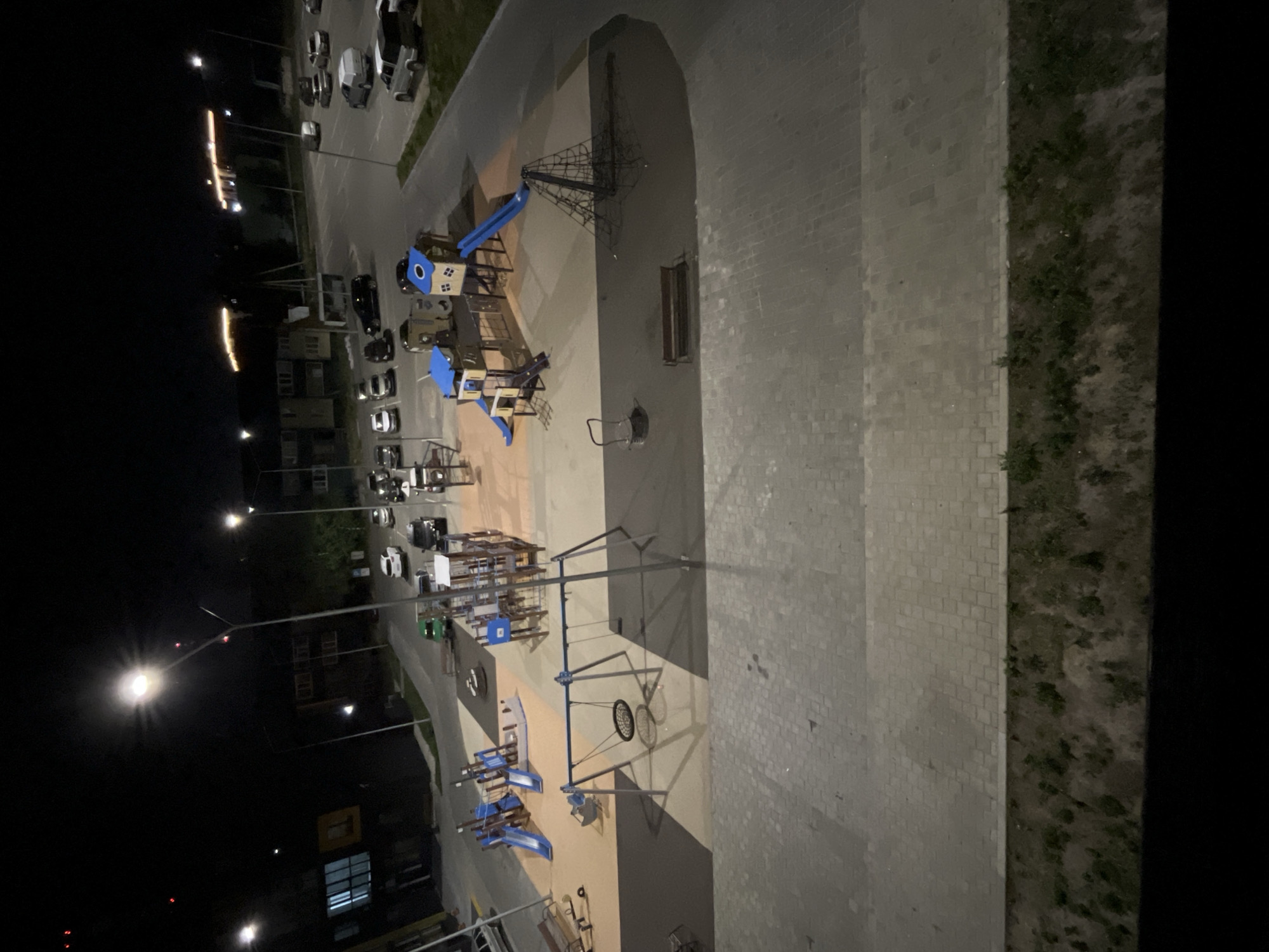 The image is an aerial view of a playground and parking lot at night in Noyabrsk, Russia. Streetlights illuminate the scene, casting long shadows. The playground features slides, swings, a climbing net, and a sandbox. Several cars are parked in the lot, and apartment buildings are visible in the background. The ground is paved with cobblestones and asphalt. The overall impression is one of urban infrastructure at rest during the night.
