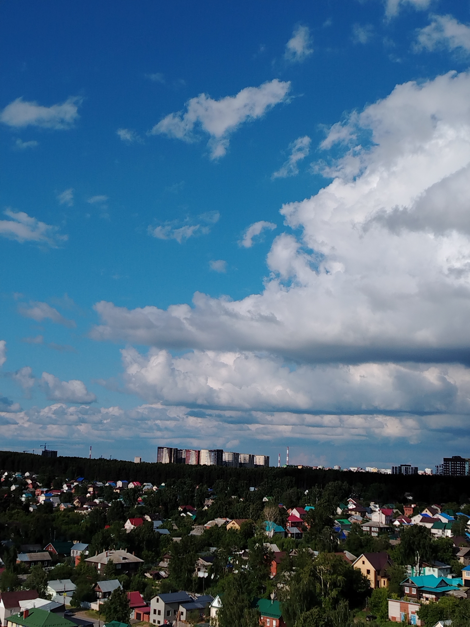 This elevated wide-angle view captures a diverse cityscape, likely Izhevsk, Russia, under a partly cloudy sky. The upper half of the image is dominated by a bright blue sky, featuring scattered white and grey cumulus clouds that vary in density, with some areas of clear blue visible.
Below the expansive sky, a dense band of dark green trees forms a significant part of the landscape. Nestled within and extending into the foreground of these trees are numerous residential houses. These homes exhibit a variety of architectural styles and a colorful array of roofs, including shades of red, green, blue, brown, and grey, suggesting a mix of older and newer structures, possibly characteristic of suburban or dacha settlements.
On the distant horizon, beyond the treeline, several clusters of modern high-rise apartment buildings are visible. Some of these multi-story structures feature bright, multi-colored facades in panels of red, white, blue, and yellow, while others appear darker and more uniform. Between these distant urban developments, two tall, slender, red-and-white striped chimneys or smokestacks are prominent, indicating an industrial presence. A faint construction crane is also discernible on the left horizon.
The scene depicts a peaceful daytime setting, bathed in clear natural light, with no explicit human activity visible. The weather is partly cloudy. No discernible text is present in the image. The overall impression is a blend of natural landscapes, traditional residential areas, and modern urban expansion.