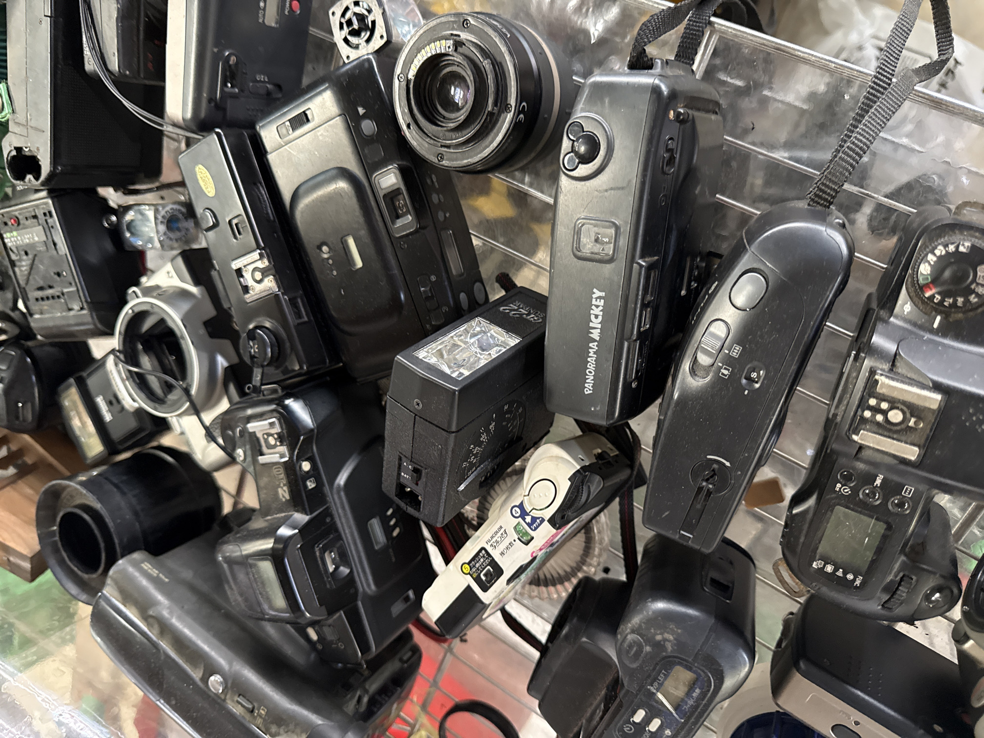 The image displays a collection of vintage cameras and camera accessories, neatly arranged on a metal shelf in what appears to be an indoor setting, possibly a retail or storage space. The scene is densely packed with various camera models, including SLR cameras, compact film cameras, and camera flashes, all in black or dark colors. A single white film camera with colorful decals stands out. In the background, hints of wooden crates and a black lens hood are visible, suggesting a workshop or a display area. The overall impression is one of a curated collection of photographic equipment. The environment is likely a shop or a collector's display area in the city of Chalong, Thailand, given the provided context, with the items resting on a reflective surface, possibly metal shelving or a display counter.