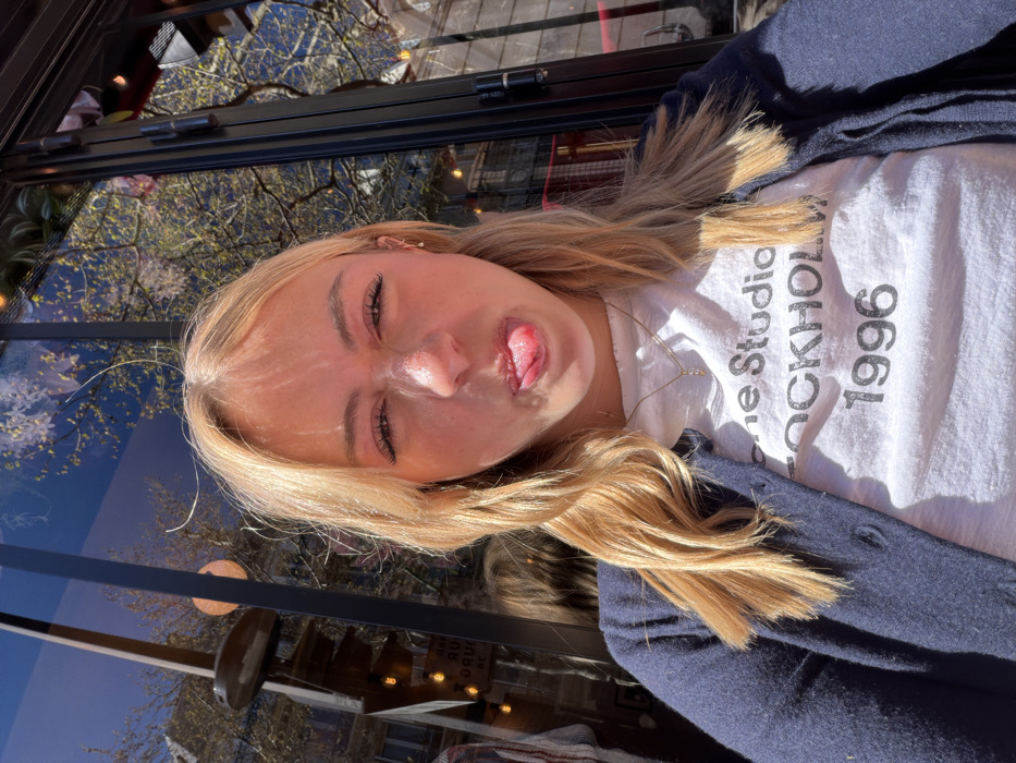 A young individual with blonde hair and fair complexion is depicted in a medium close-up, making a playful facial expression by scrunching their nose and extending their tongue. They are wearing a white t-shirt with "Acne Studios STOCKHOLM 1996" printed in black text, and a navy blue button-up cardigan over it. A delicate gold-colored necklace with a small, curved pendant is visible. The background consists of dark, reflective glass surfaces, likely storefront windows or a cafe facade. These surfaces show clear reflections of an outdoor urban environment, including traditional light-colored stone buildings characteristic of Parisian architecture, trees with sparse foliage under a clear blue sky, and spherical orange light fixtures. Reversed French text, including "AUX DOUCEURS DE" and fragments suggestive of a "BOULANGERIE," is discernible in the reflections, indicating the scene is located on a street in Paris, France.