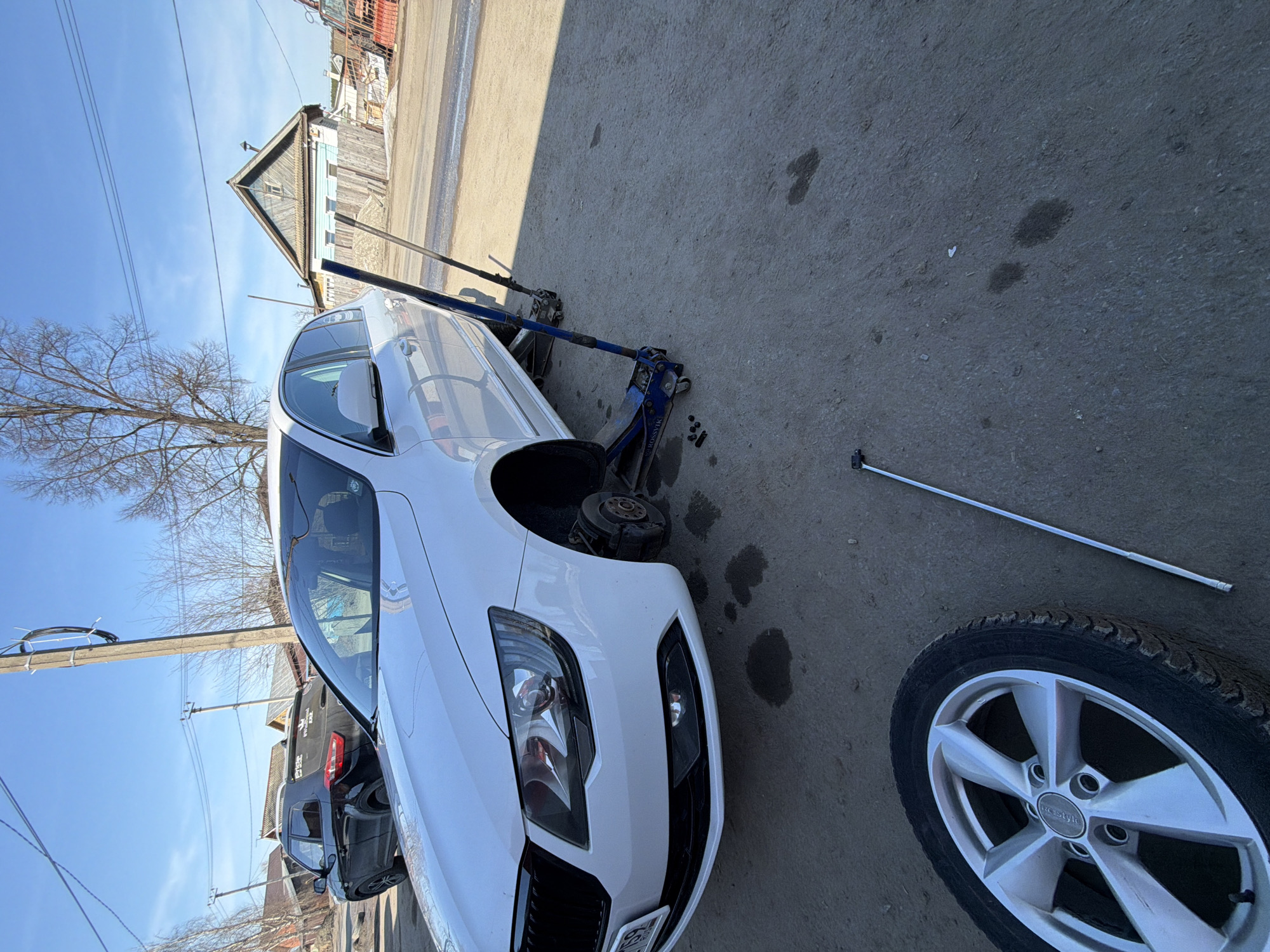 A white car is elevated on two blue jacks, its front driver's side wheel removed and lying nearby. A lug wrench rests on the ground. The scene appears to be outdoors on a paved surface with some oil stains. In the background are buildings and a clear blue sky. Based on visual cues, this image could be taken in a roadside setting. There is no indication as to whether this photo was taken in the city of Chaikovsky, Russia.